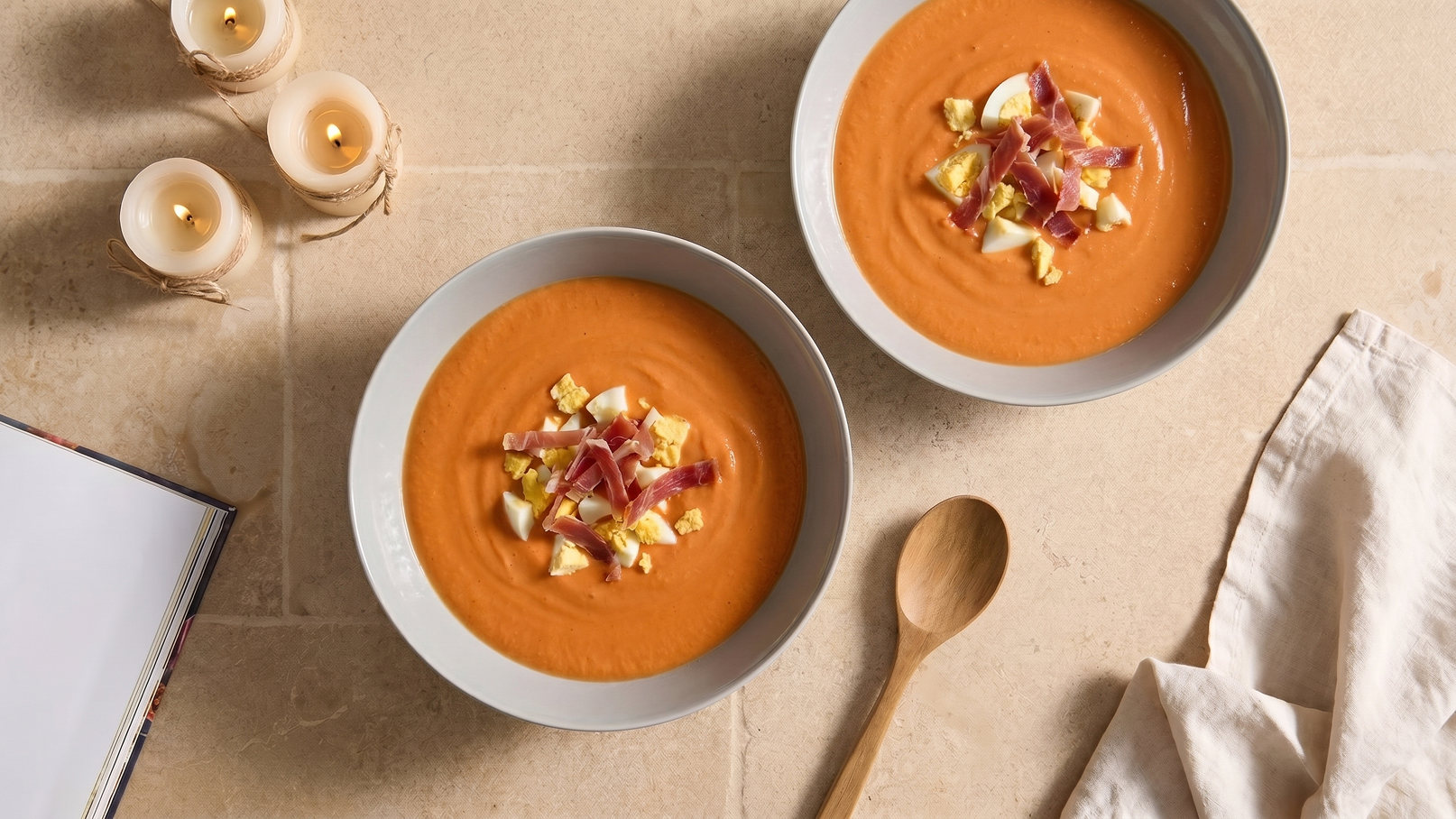 Zwei Schüsseln mit samtiger Tomatensuppe stehen auf einem Tisch. Die Suppe ist mit gehacktem Ei und Schinken dekoriert. Neben den Schüsseln liegen ein Holzlöffel und ein geöffnetes Kochbuch, während einige Kerzen im Hintergrund brennen.