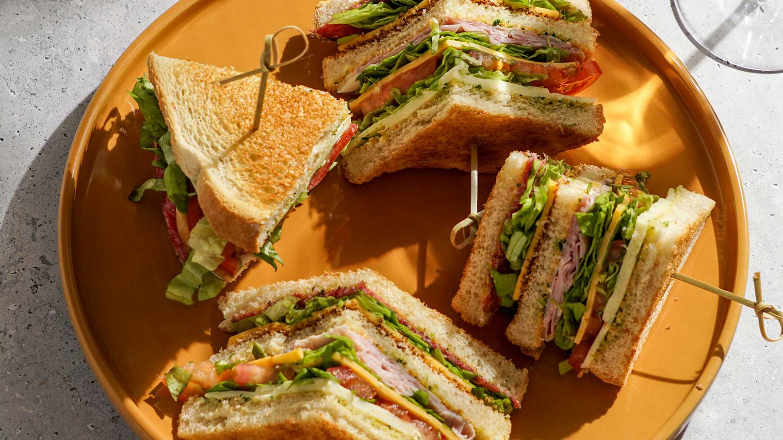 Ein Teller mit mehreren Sandwiches, die in Stücke geschnitten sind. Die Sandwiches sind mit verschiedenen Zutaten gefüllt, darunter Salat, Tomaten und Aufschnitt. Die Brotscheiben sind knusprig gebräunt. Ein Zahnstocher hält einige der Stücke zusammen.