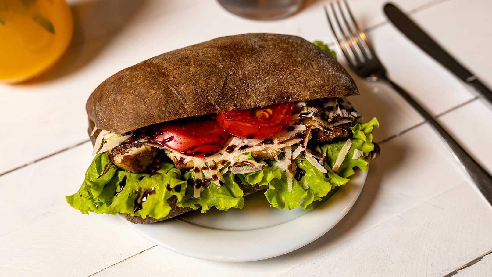 Ein Sandwich mit einem dunklen Brötchen, gefüllt mit gegrilltem Fleisch, frischen Tomatenscheiben, geriebenem Käse und knackigem Salat. Es steht auf einem weißen Teller, umgeben von Besteck.