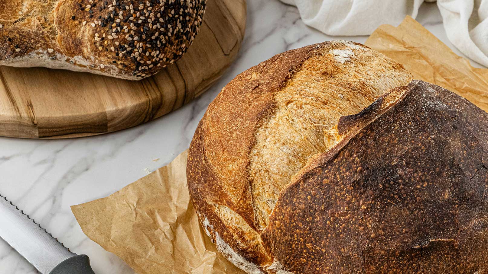 Im Bild sind zwei Brote auf einem Holzbrett und einem Stück Papier zu sehen. Eines der Brote hat eine knusprige, goldbraune Kruste, während das andere mit verschiedenen Samen bestreut ist. Ein Brotmesser liegt daneben. Die Szene vermittelt ein Gefühl von frisch gebackenem Brot.