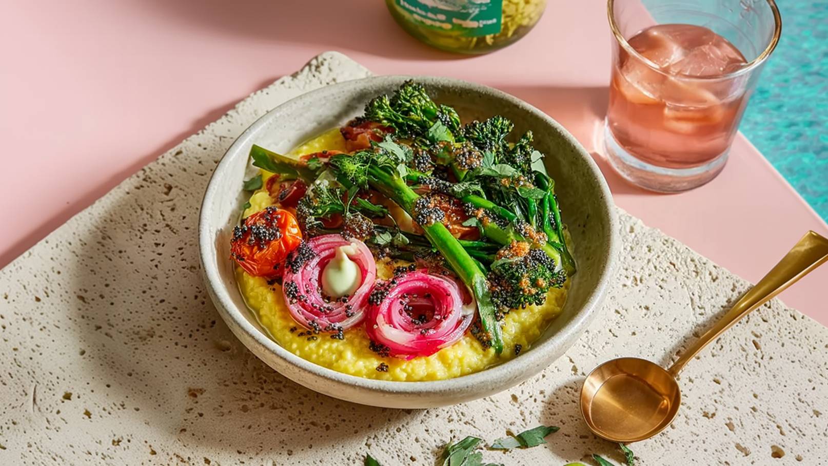 Eine Schüssel mit cremigem Polenta-Gericht, garniert mit gebratenem Gemüse wie Brokkoli, Kirschtomaten und roten Zwiebeln. Ein Hauch von Kräutern und Gewürzen vollendet das Bild. Im Hintergrund ist ein Glas mit Getränk und ein goldener Löffel zu sehen.