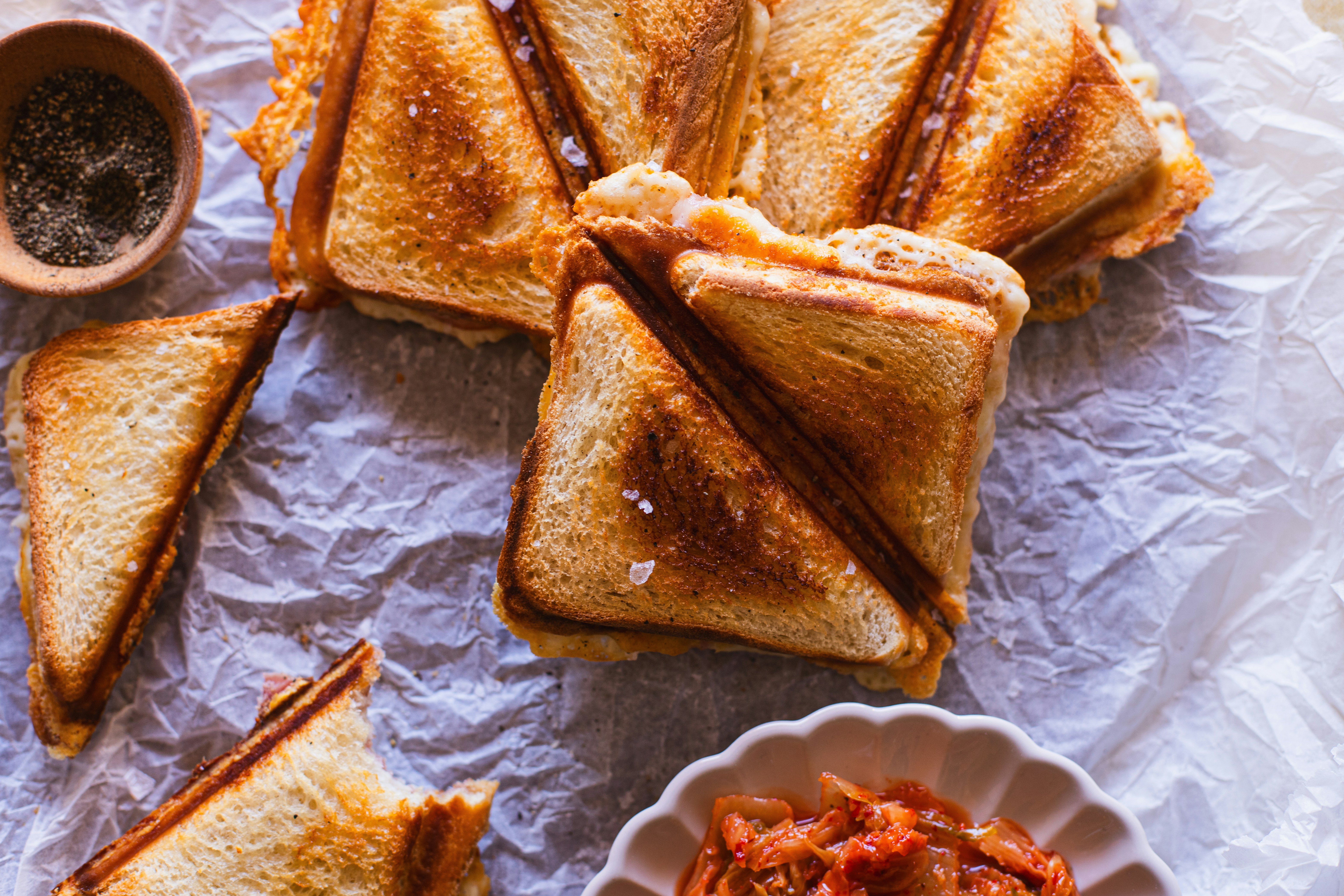 Das Bild zeigt mehrere goldbraun geröstete Sandwiches, die diagonal geschnitten sind und auf Pergamentpapier liegen. Daneben befindet sich eine kleine Schüssel mit einer würzigen roten Beilage. Die Sandwiches wirken knusprig und appetitlich.