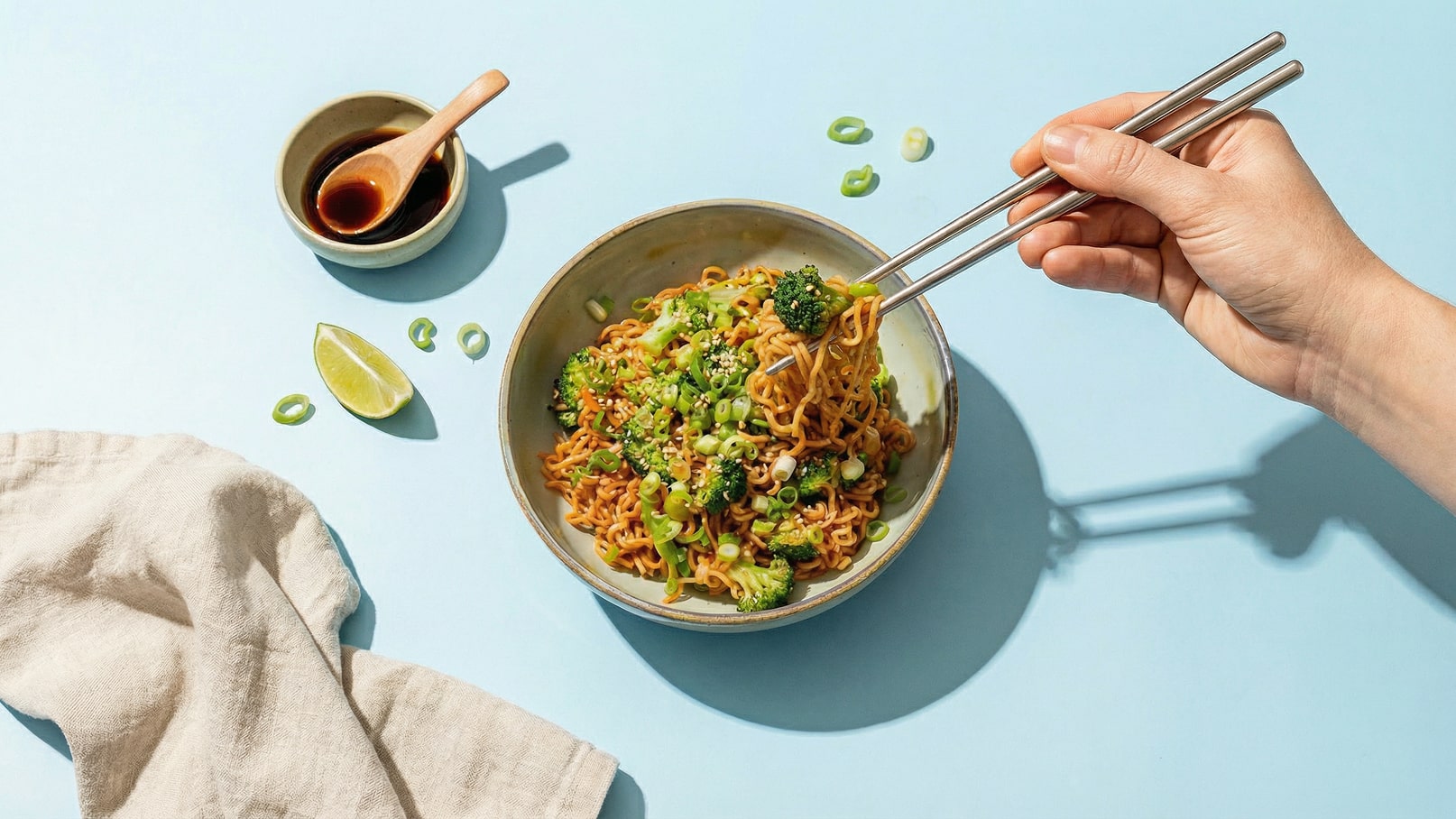 Eine Hand hält Essstäbchen über einer Schüssel mit gebratenen Nudeln, garniert mit Brokkoli und Frühlingszwiebeln. Im Hintergrund befindet sich eine kleine Schale mit Sojasauce und ein Limettenstück. Der Hintergrund ist hellblau.