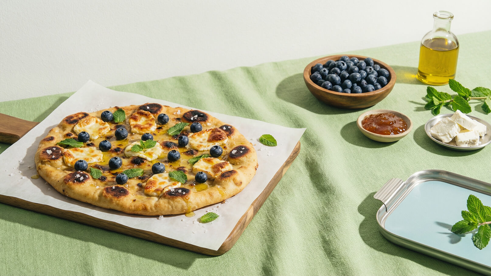 Ein frisch gebackener Pizzaboden mit geschmolzenem Käse, reich garniert mit Heidelbeeren und frischen Minzblättern. Daneben ein Holzbrett, eine Schale mit weiteren Heidelbeeren, etwas Olivenöl, Marmelade und Käse. Der Hintergrund ist in sanften Grüntönen gehalten.