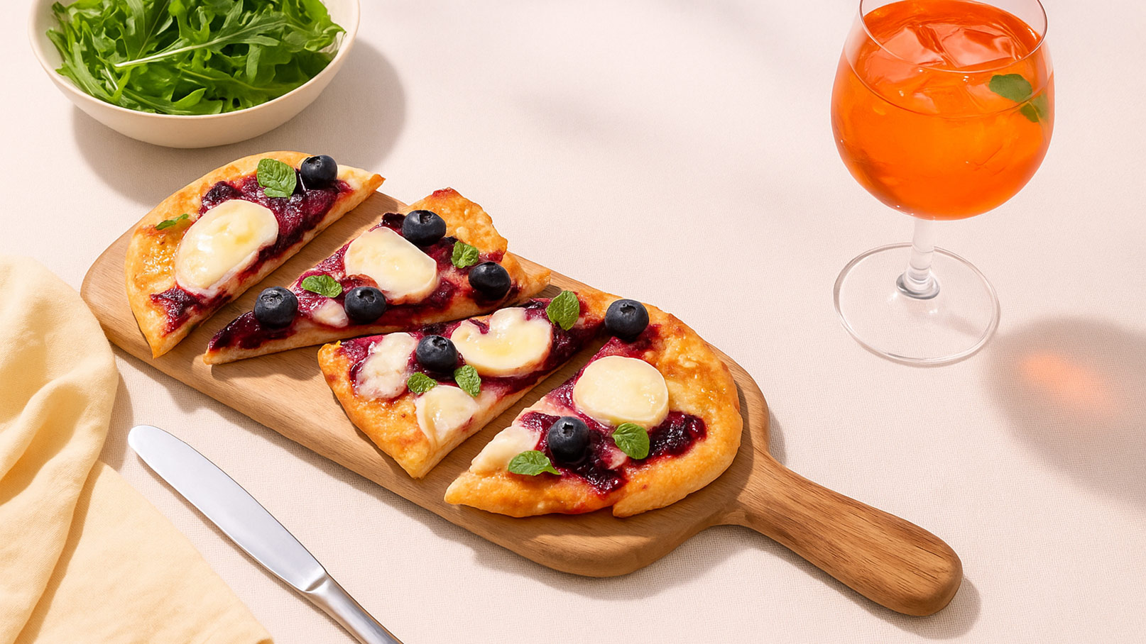 Ein Holzbrett mit zwei Pizzastücken, belegt mit Blaubeeren und Schmelzkäse, liegt auf einem Tisch. Daneben steht ein elegantes Glas mit einem orangen Getränk. Im Hintergrund liegt eine Schüssel mit frischem Rucola.