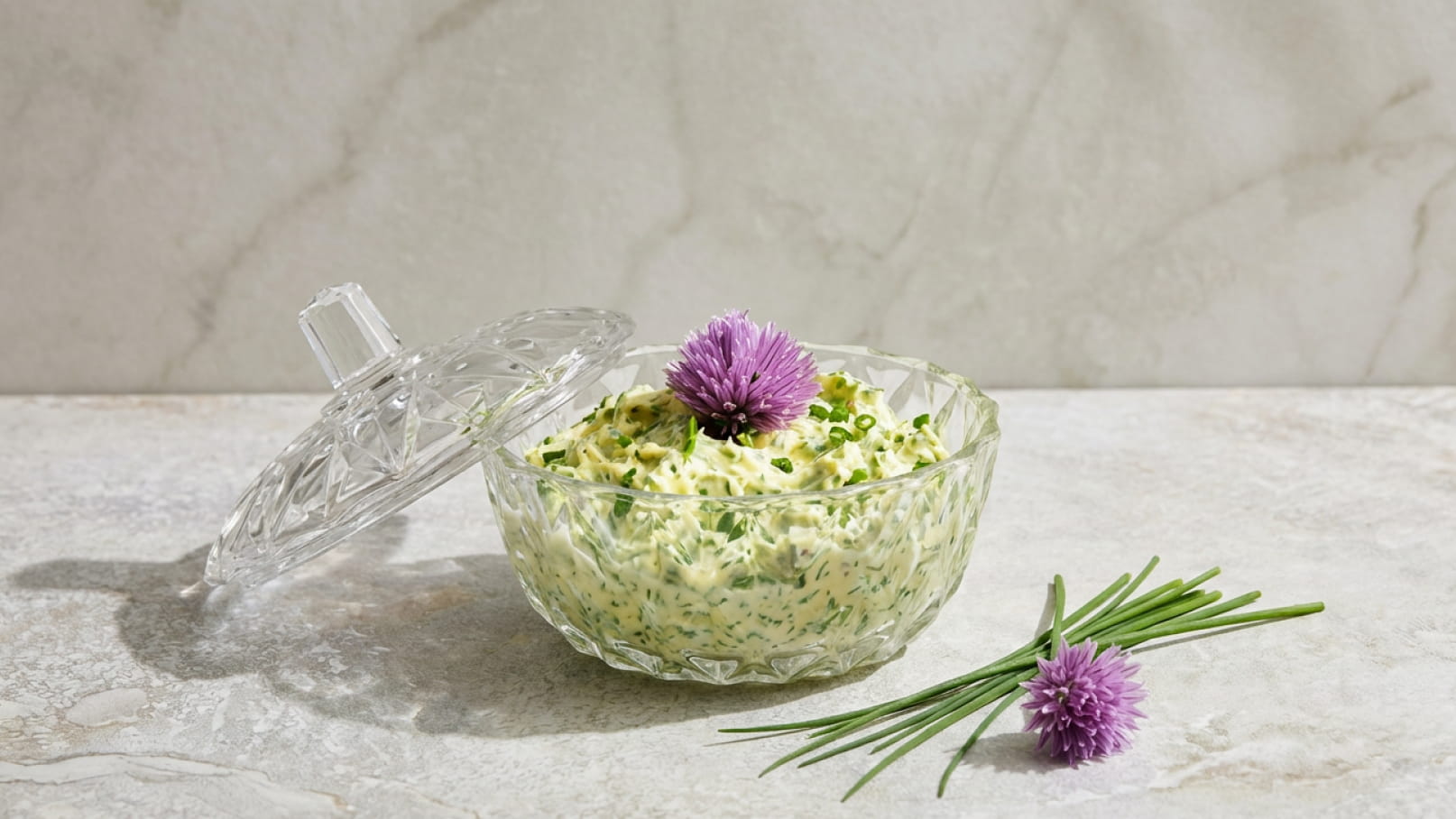 Eine gläserne Schüssel mit frischem Schnittlauch-Dip, dekoriert mit einer lila Blüte, steht auf einer hellen Oberfläche. Daneben liegen einige Schnittlauchzweige. Der Deckel der Schüssel ist leicht abgehoben.