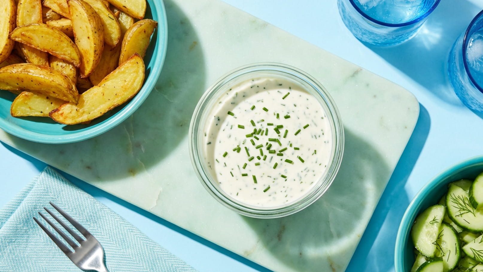 Auf einem hellblauen Tisch liegen knusprige Kartoffelspalten in einer türkisfarbenen Schüssel. Daneben befindet sich eine Schüssel mit einer cremigen Sauce, die mit Schnittlauch garniert ist. Im Hintergrund sind Gläser mit Wasser und ein kleiner Teller mit Gurkenscheiben zu sehen.