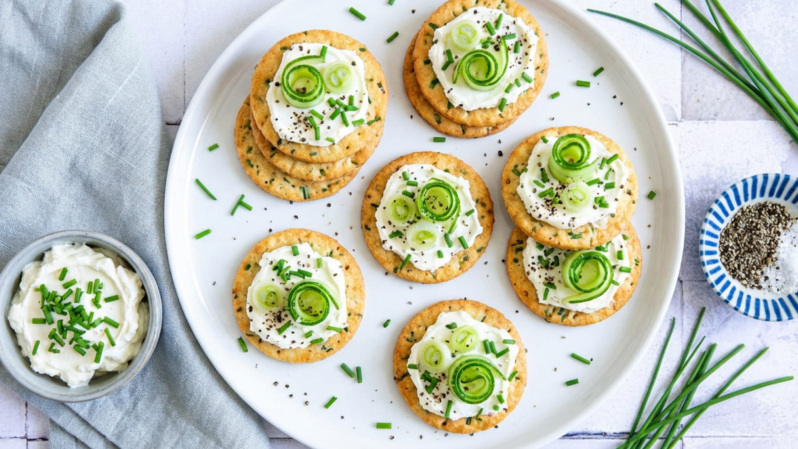 Auf einem weißen Teller liegen mehrere Cracker mit Frischkäse. Die Cracker sind mit dünn geschnittenen Frühlingszwiebeln und Schnittlauch garniert. Neben dem Teller steht eine Schüssel mit cremigem Frischkäse und etwas gehacktem Schnittlauch.