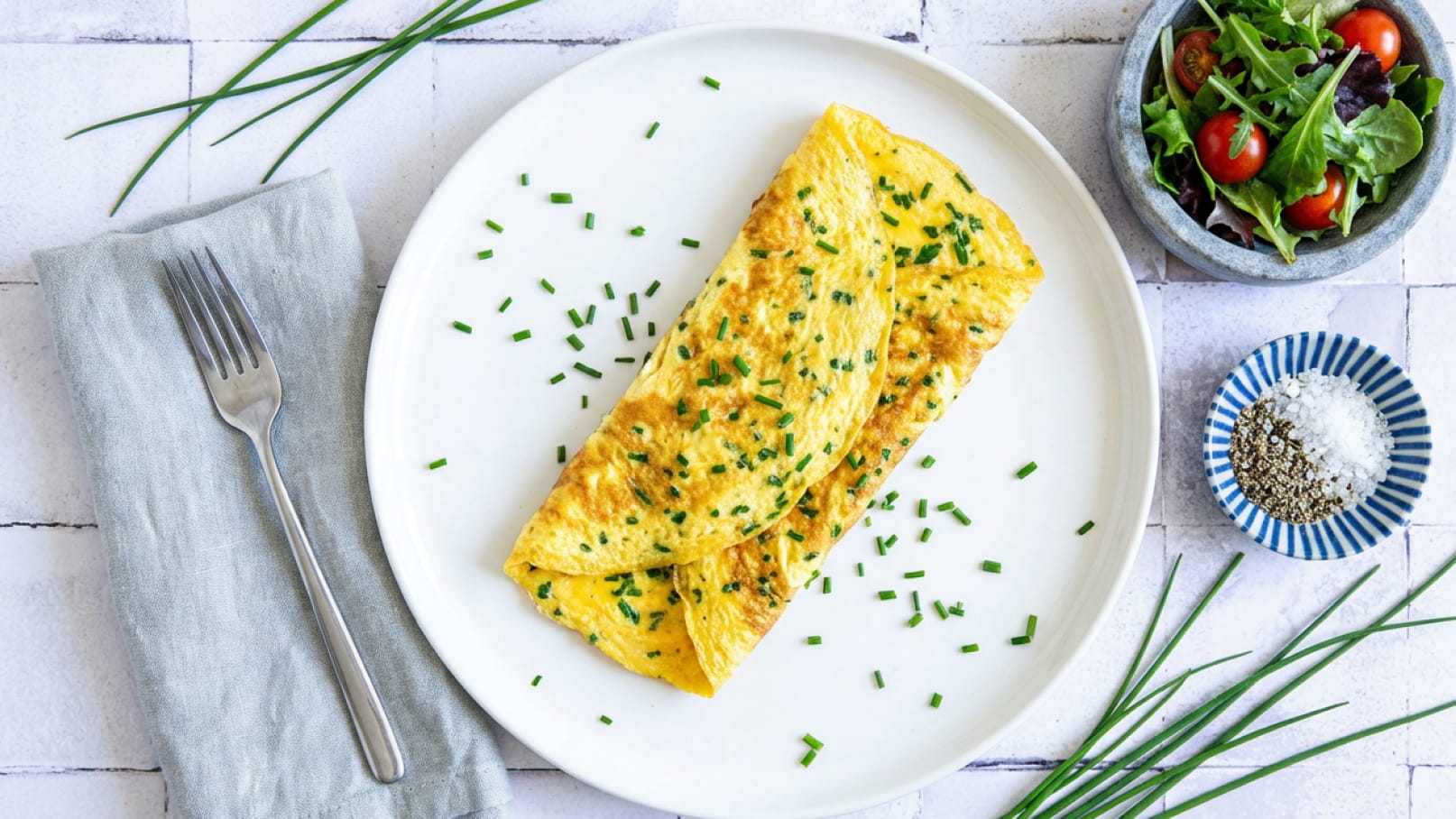 Ein Teller mit einem goldbraunen Omelett, das mit Schnittlauch bestreut ist. Neben dem Teller liegt eine Gabel auf einer grauen Serviette. Im Hintergrund befindet sich eine Schüssel mit frischem Salat und Kirschtomaten.
