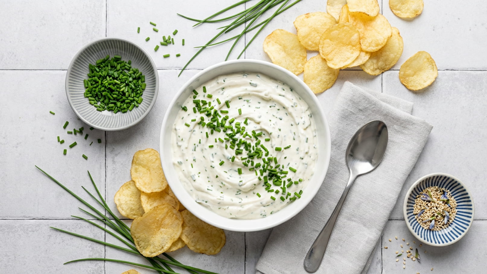Eine weiße Schüssel mit cremigem Dip, garniert mit frischem Schnittlauch. Um die Schüssel herum liegen knusprige Kartoffelchips, eine kleine Schale mit gehacktem Schnittlauch und eine weitere Schale mit Gewürzen. Ein Löffel liegt neben der Schüssel auf einem weißen Servietten.