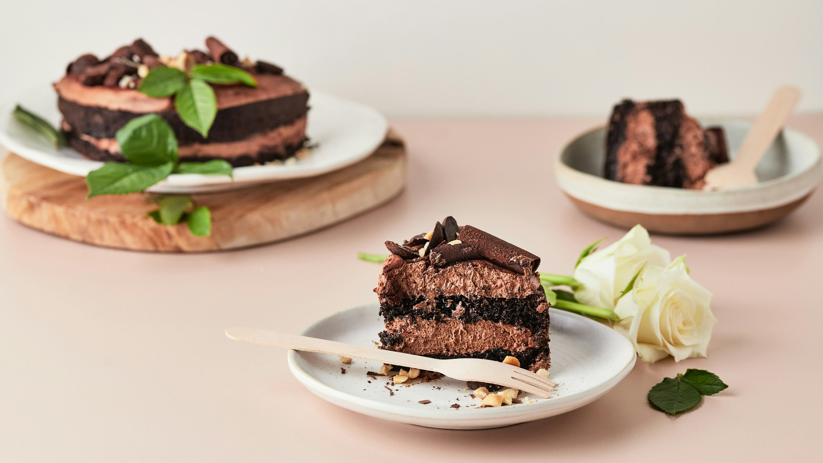 Ein Stück Schokoladentorte steht auf einem weißen Teller, umgeben von Walnüssen und Schokoladenraspeln. Im Hintergrund sind eine ganze Torte und ein weiterer Teller mit Tortenstück sichtbar. Einige grüne Blätter und eine weiße Rose ergänzen die Szene.