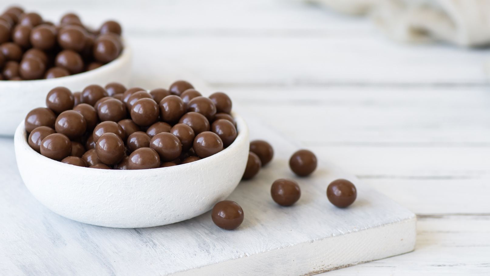 Zwei weiße Schalen auf einem Holzbrett sind mit runden, glänzenden, schokoladenbraunen Bonbons gefüllt. Einige der Bonbons liegen locker auf dem Brett verteilt. Die Szene ist hell und minimalistisch gestaltet.
