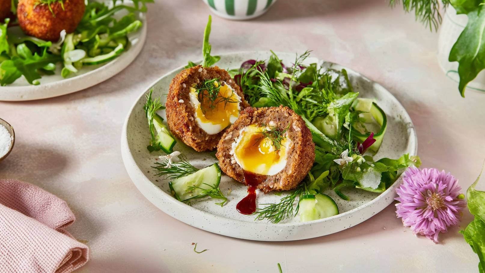 Auf dem Bild sind zwei aufgeschnittene, frittierte Eierbällchen mit flüssigem Eigelb zu sehen, serviert auf einem Teller mit frischem Salat und Gurkenscheiben. Das Gericht ist mit frischen Kräutern garniert.
