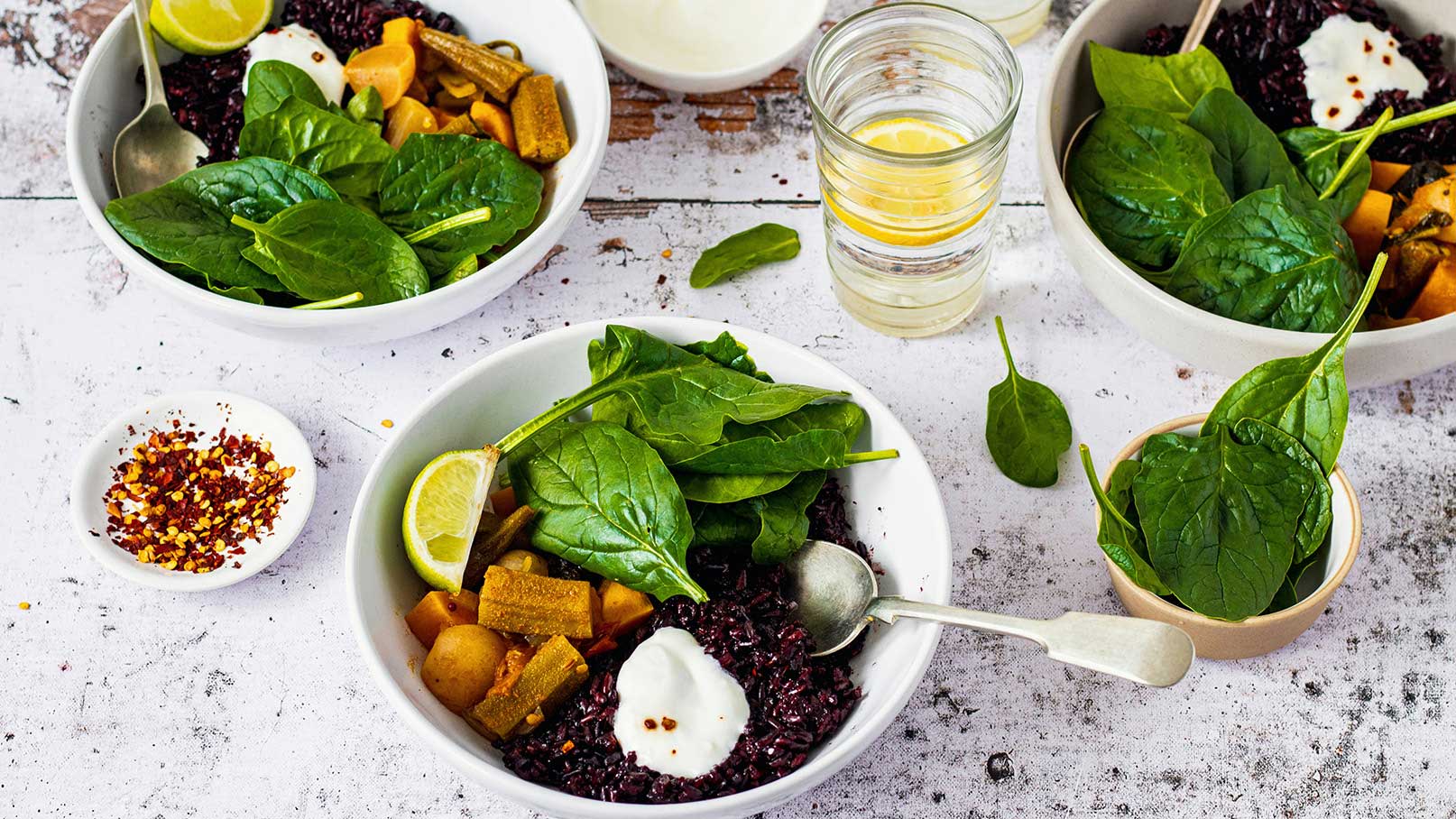 Eine Schüssel mit schwarzem Reis, frischem Spinat, Wurzelgemüse und einem Klecks Joghurt. Neben der Schüssel liegen eine Limettenscheibe und eine kleine Schale mit Chiliflocken. Im Hintergrund ist ein Glas Wasser mit Zitronenscheiben zu sehen.
