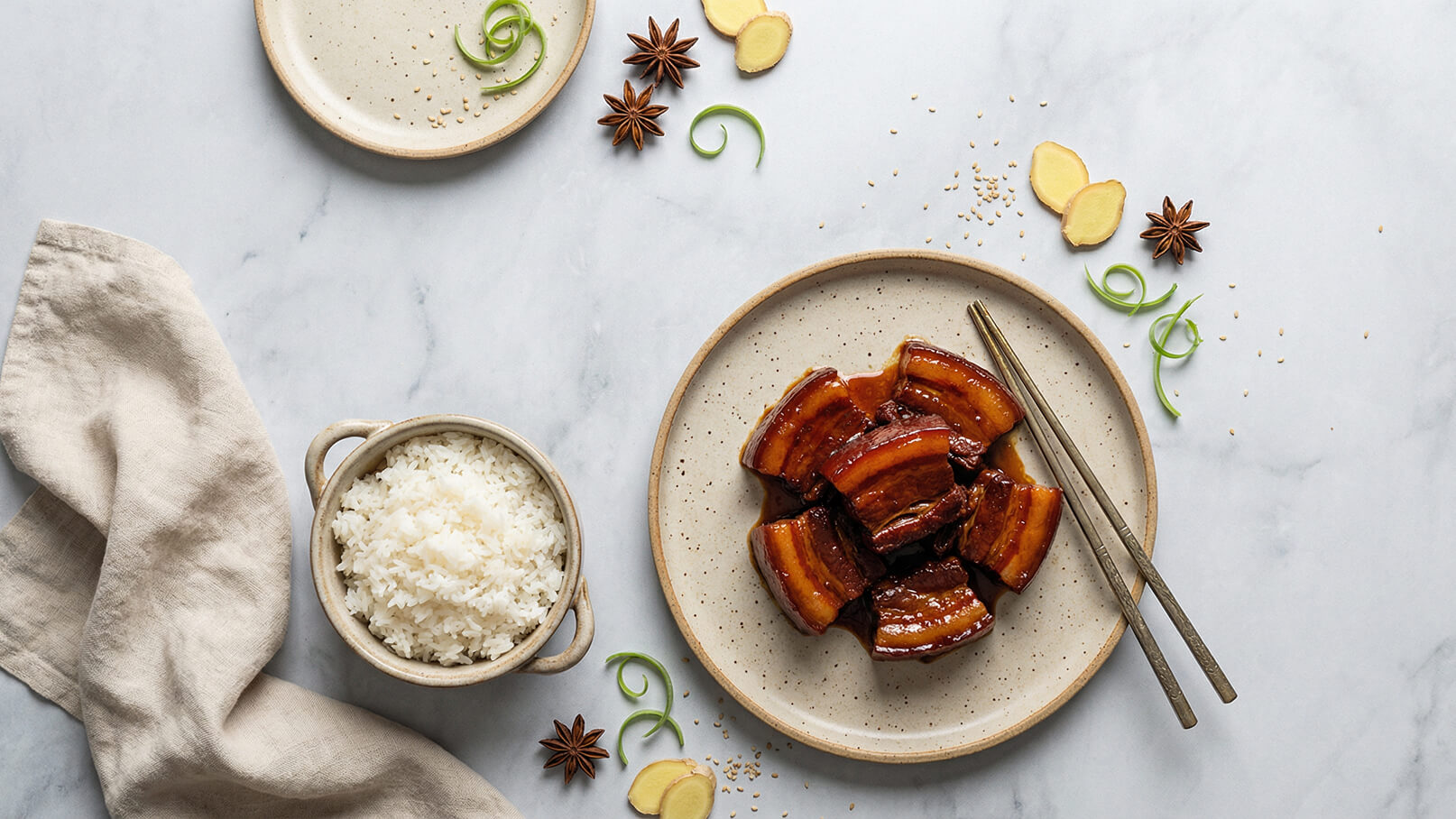 Ein Teller mit saftigem, glasiertem Fleisch ist in der Mitte der Abbildung angeordnet. Daneben befindet sich eine Schüssel mit gedämpftem Reis. Gewürze wie Sternanis und frischer Ingwer sind verstreut, während einige Frühlingszwiebeln als Garnitur dienen.
