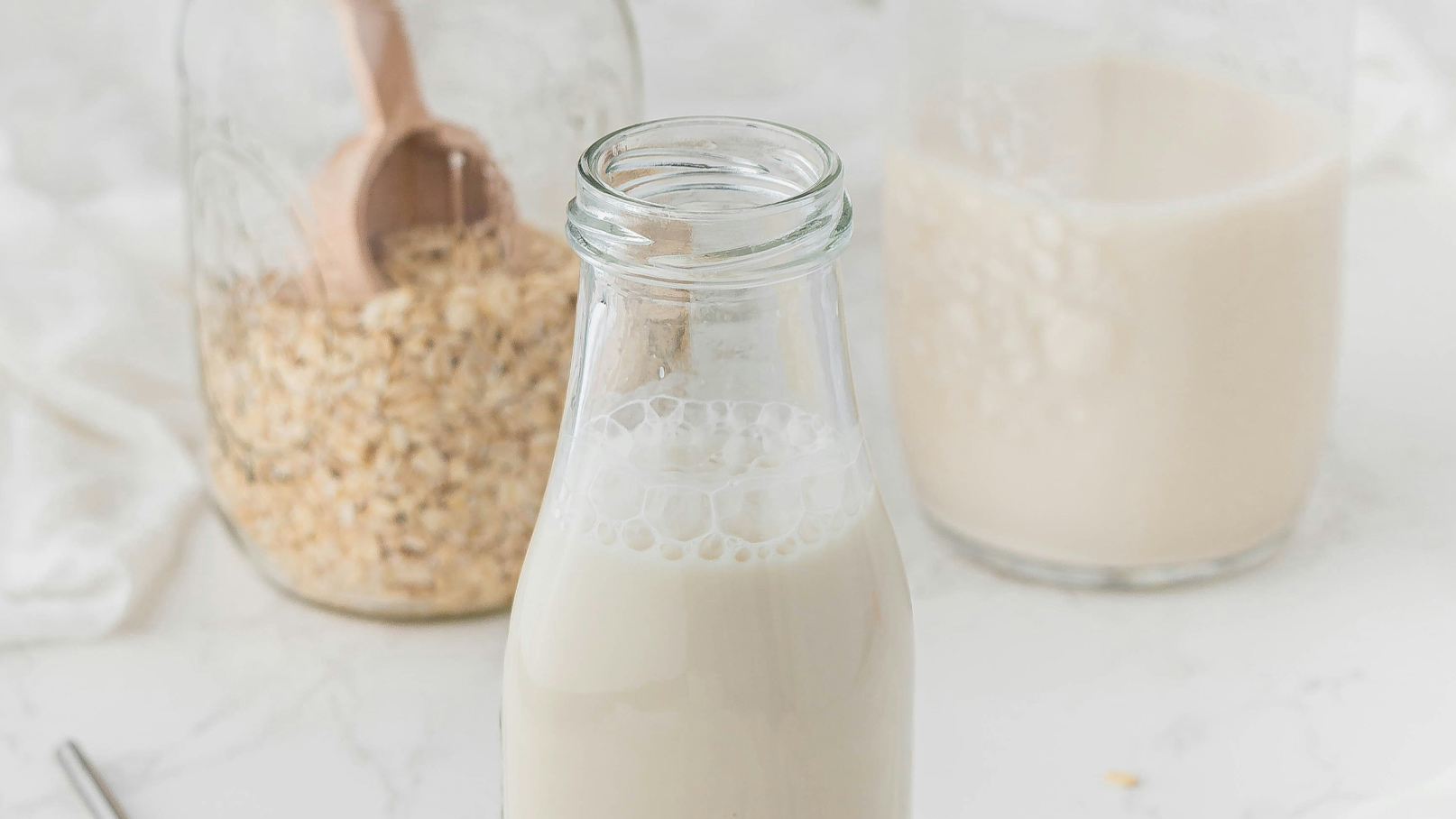 Eine Glasflasche mit frischer Milch steht im Vordergrund. Im Hintergrund sind ein Behälter mit Haferflocken und ein weiterer Behälter mit einer cremigen Flüssigkeit zu sehen. Die Szene vermittelt eine gemütliche Küchensituation.