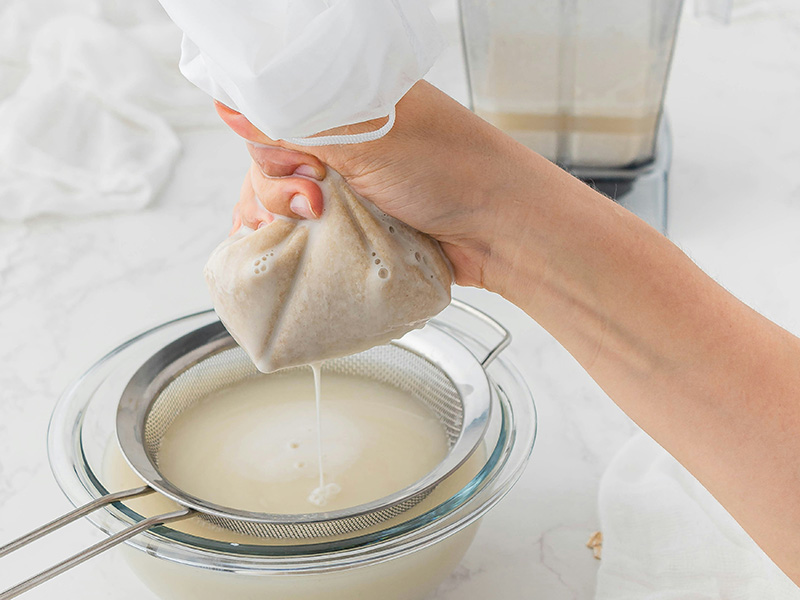 Eine Hand hält einen Beutel mit einer cremigen Substanz über ein Sieb, aus dem Flüssigkeit in eine Schüssel tropft. Im Hintergrund ist ein Mixer sichtbar. Die Szene zeigt den Prozess des Abseihens, möglicherweise zur Herstellung pflanzlicher Milch.