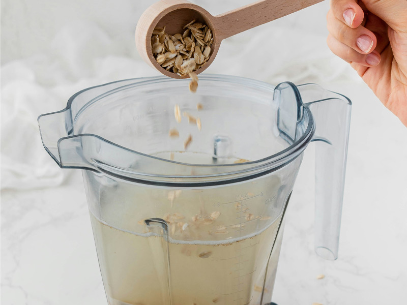 Eine Hand hält einen Holzlöffel mit Haferflocken über einen Mixer, in den die Flocken gerade hineingestreut werden. Der Mixer enthält eine Flüssigkeit, die klar und teilweise durchsichtig ist.