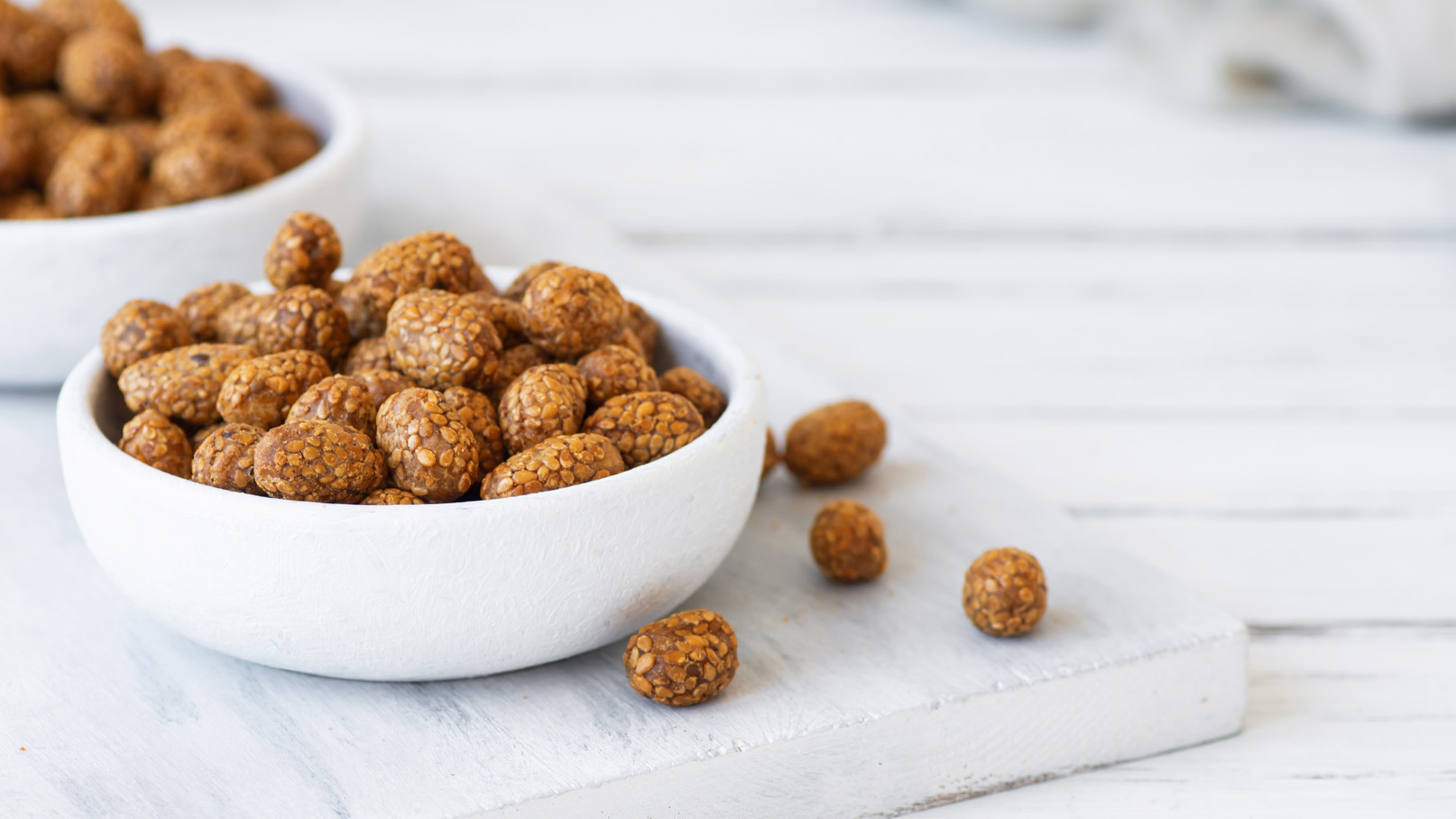 Eine weiße Schüssel enthält zahlreiche kleine, runde Snacks, die eine knusprige Oberfläche und eine braune Farbe haben. Einige dieser Snacks liegen verstreut auf einem weißen Untergrund. Im Hintergrund ist eine weitere Schüssel mit ähnlichen Snacks zu sehen.