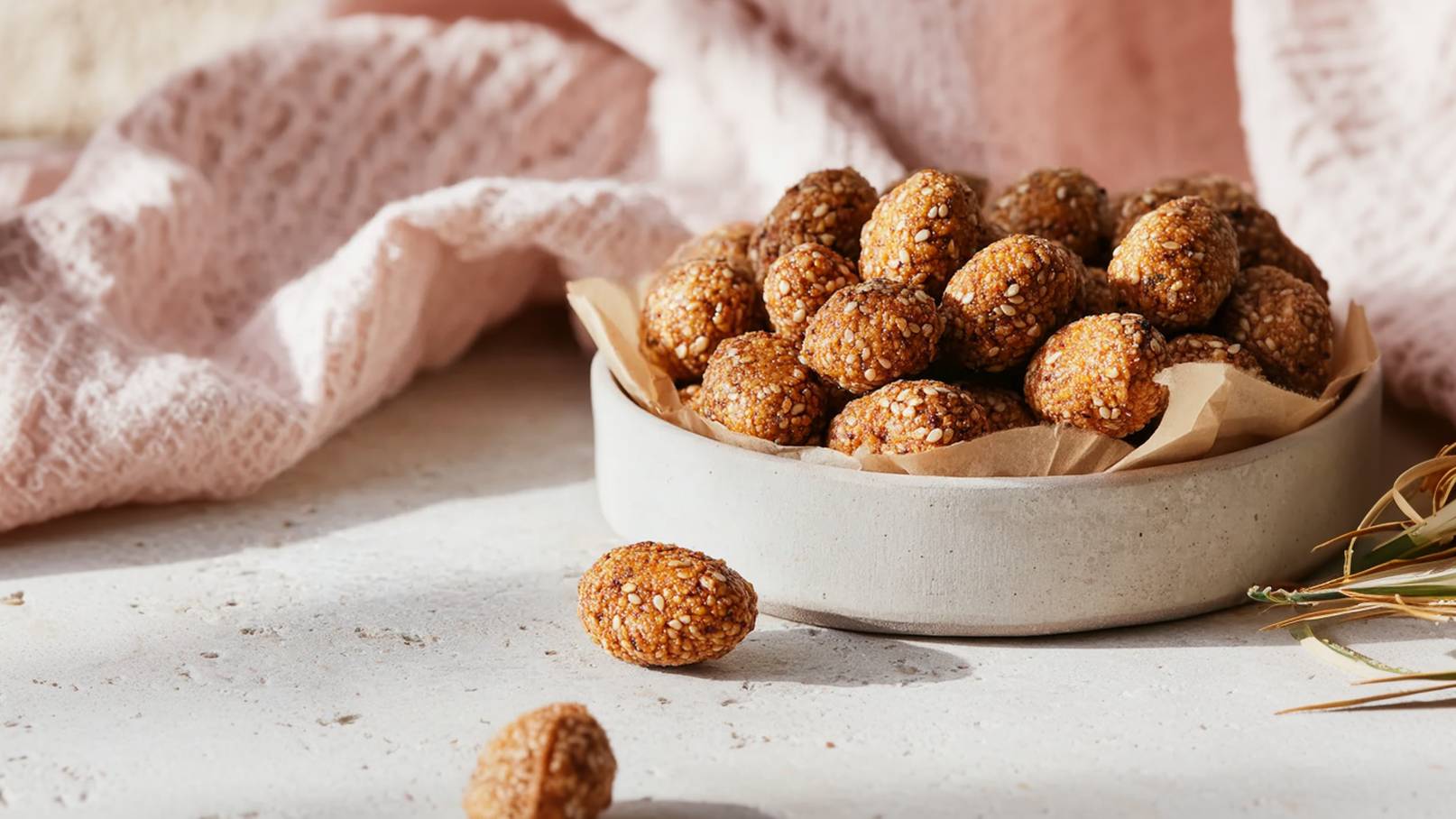 Eine weiße Schüssel enthält zahlreiche kleine, runde Snacks, die eine knusprige Oberfläche und eine braune Farbe haben. Einige dieser Snacks liegen verstreut auf einem weißen Untergrund. Im Hintergrund ist eine weitere Schüssel mit ähnlichen Snacks zu sehen.