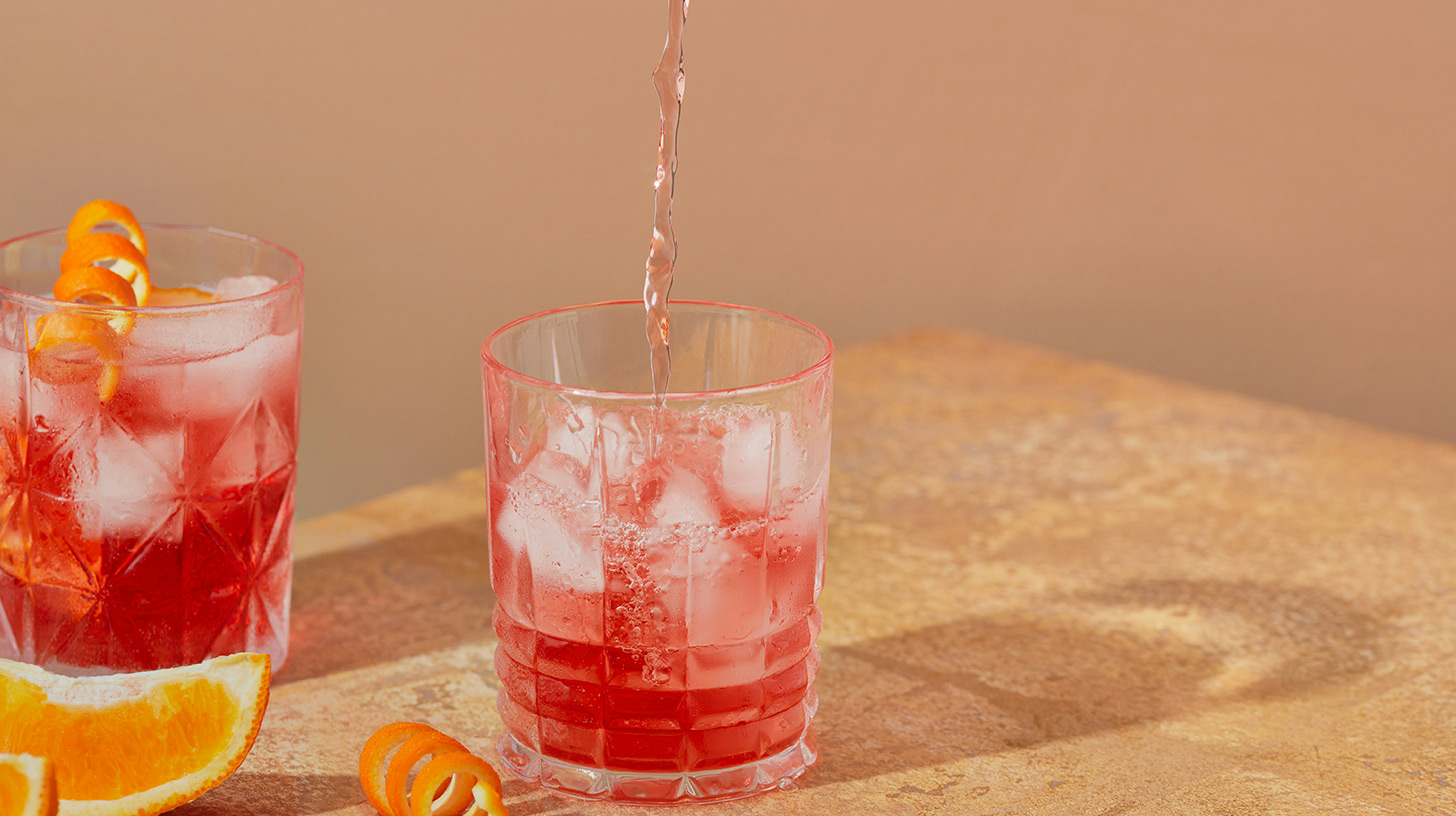 Zwei Gläser mit einem roten Getränk, gefüllt mit Eiswürfeln. Eines der Gläser wird gerade nachgefüllt. Auf dem Tisch liegen Orangenscheiben und eine Spiralzeste. Der Hintergrund ist beige.