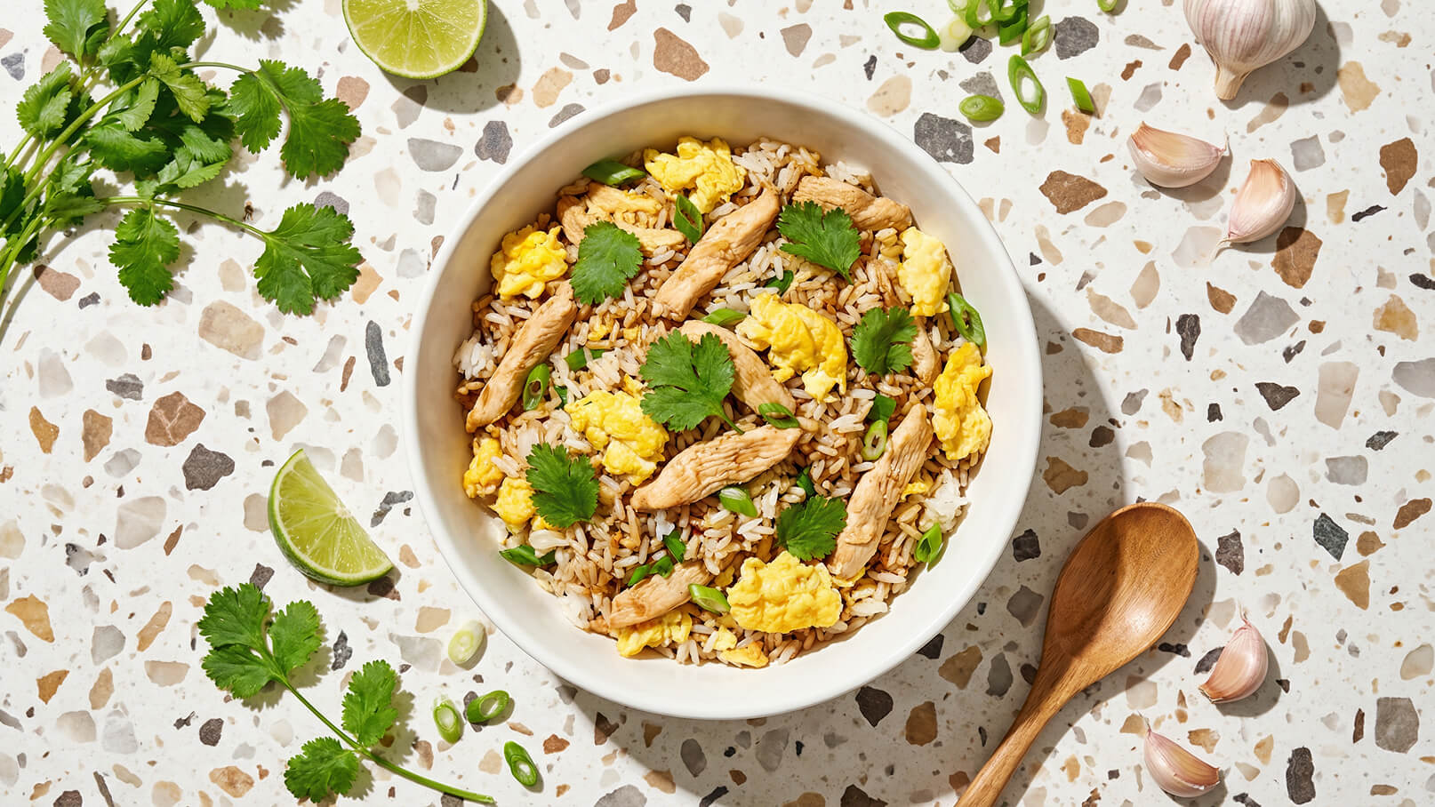 Eine Schüssel mit gebratenem Reis, garniert mit zarten Hühnchenstreifen, Rührei, frischem Koriander und Frühlingszwiebeln. Neben der Schüssel liegen Limettenscheiben und einige Knoblauchzehen. Der Hintergrund ist eine helle, strukturierte Oberfläche.