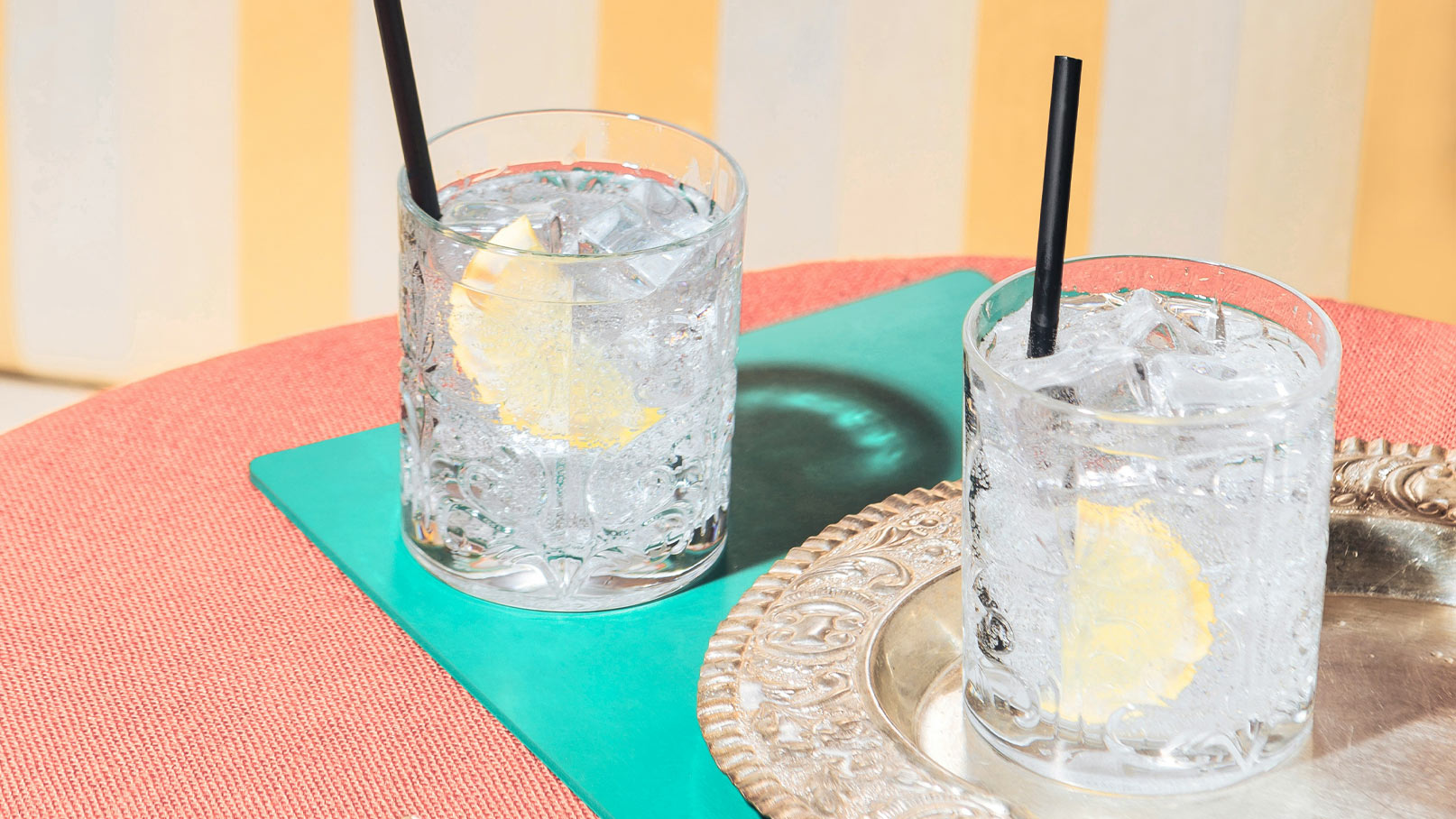 Zwei Gläser mit Eiswasser und Zitronenscheiben stehen auf einem Tisch. Eines der Gläser hat einen schwarzen Strohhalm. Die Gläser sind auf einer grünen Unterlage, die auf einem roten Tisch liegt. Im Hintergrund sind gestreifte Wände zu sehen.