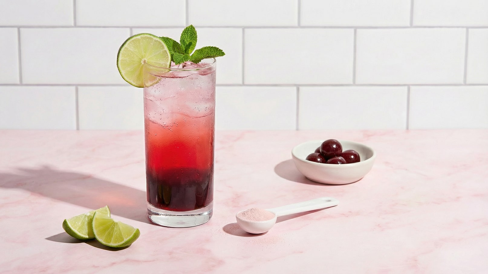 Ein hoher Cocktailgläser gefüllt mit einem mehrschichtigen Getränk, das rot und pink aussieht, garniert mit einem Limettenschnitz und frischen Minzblättern. Daneben liegen halbierte Limetten, eine kleine Schaufel mit rosa Pulver und eine Schale mit dunklen Kirschen.