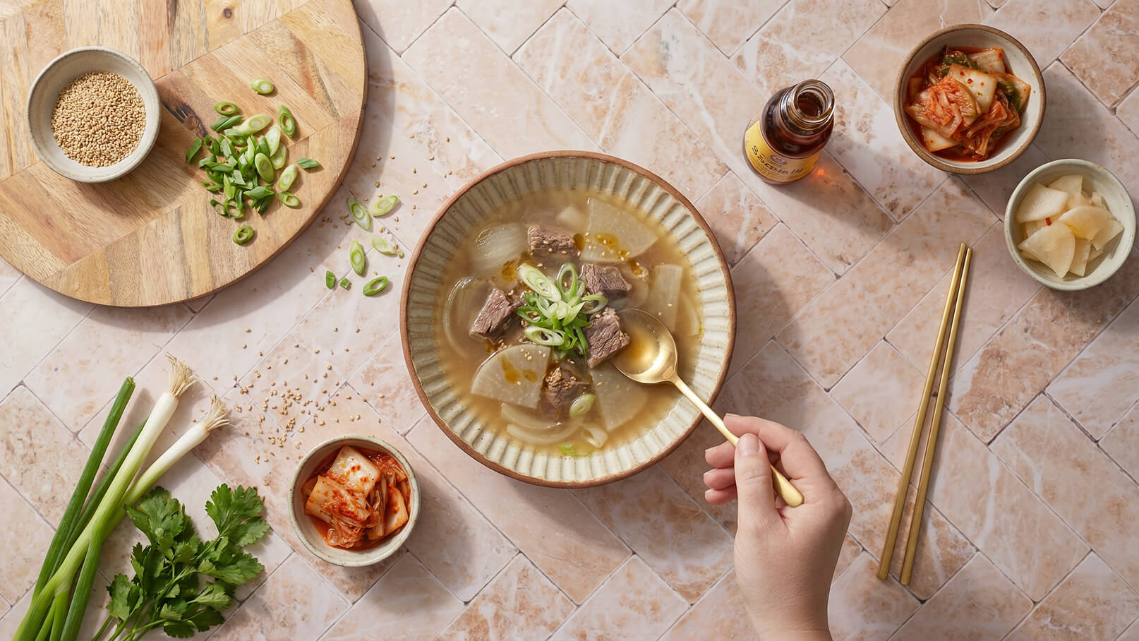 Eine Schüssel Suppe mit Stückchen Fleisch und Gemüse, garniert mit Frühlingszwiebeln und Sesam. Neben der Suppe sind zwei kleine Schalen mit Kimchi und eingelegtem Gemüse sowie eine Flasche Sojasauce. Eine Hand mit einem Löffel greift nach der Suppe.