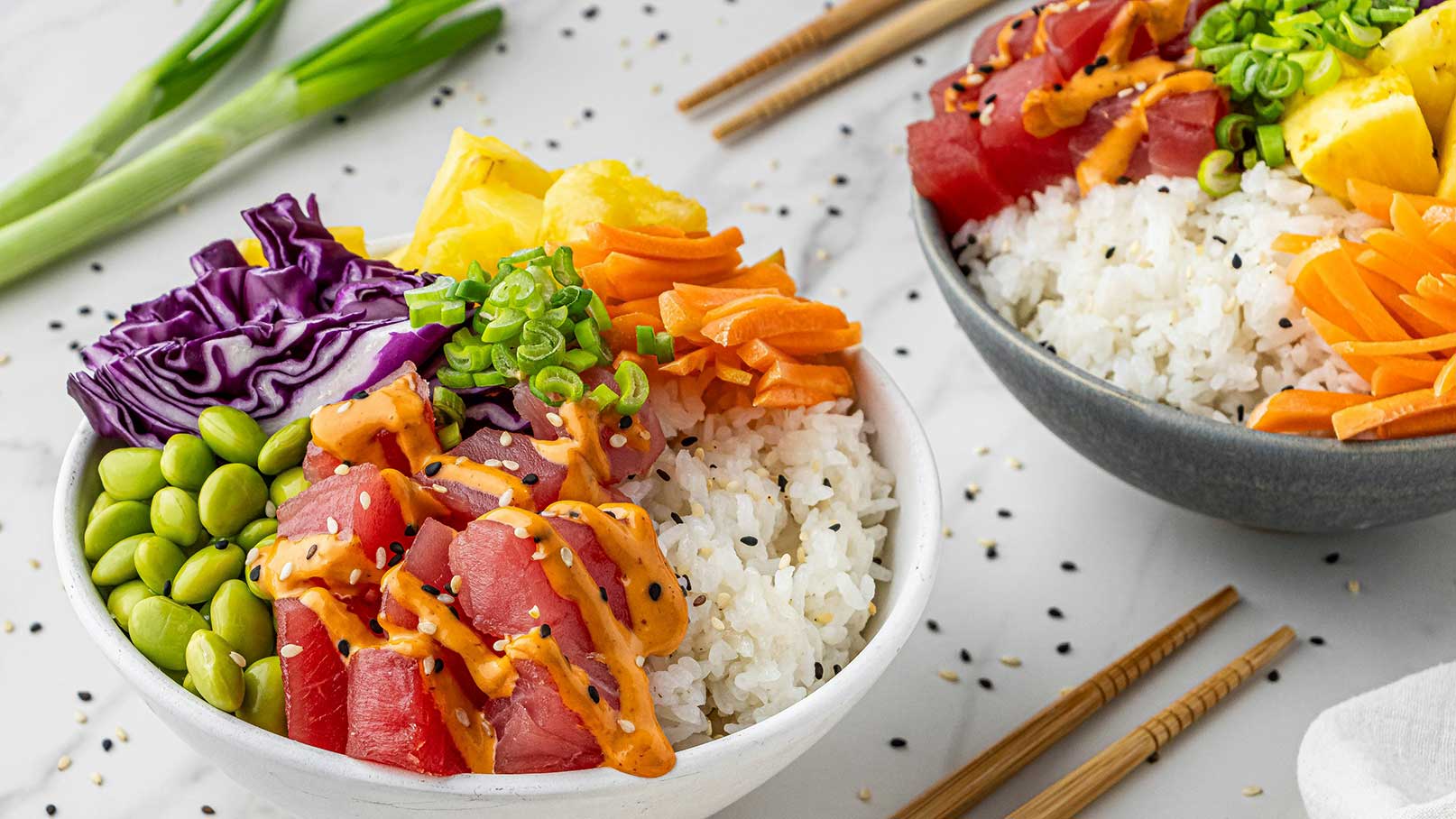 Eine Schüssel mit Sushi-Reis, belegt mit frischem Thunfisch, buntem Gemüse wie lila Kohl, Karotten und Fruchtstücken, garniert mit grünen Zwiebeln und einer würzigen Soße. Neben der Schüssel liegen Essstäbchen auf einem hellen Untergrund.
