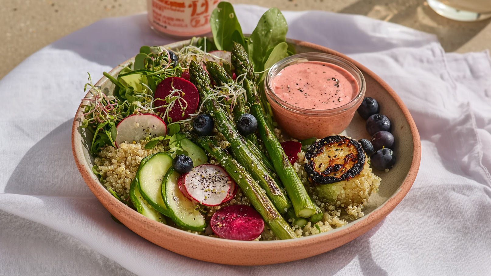 Eine Schüssel mit frischem Salat, bestehend aus Spargel, Radieschen, Gurken und Quinoa. Neben dem Salat liegt eine kleine Schale mit einer rosa Soße sowie einige Heidelbeeren. Der Teller befindet sich auf einem weißen Tuch.