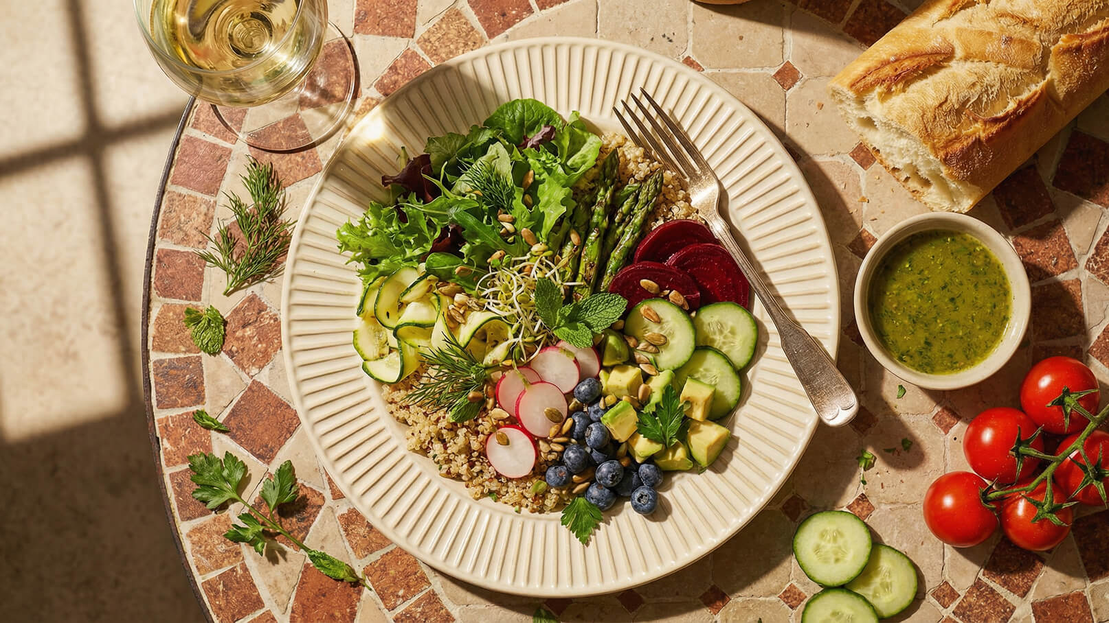 Ein Teller mit einer bunten Salatmischung aus Blattsalaten, Gurken, Radieschen, Avocado, Quinoa und Sprossen, garniert mit Sonnenblumenkernen. Neben dem Teller befinden sich ein Glas Weißwein, ein Schälchen mit grüner Sauce und frische Kirschtomaten.