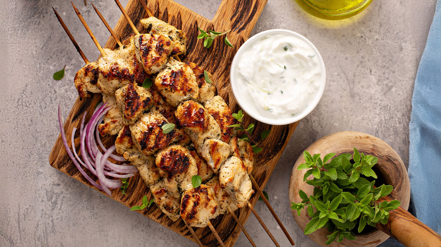 Gegrillte Hähnchenspieße liegen auf einem Holzbrett, garniert mit frischen Kräutern. Daneben befindet sich eine kleine Schale mit Dip, vermutlich Joghurtsauce. Ein paar rote Zwiebelringe sind ebenfalls sichtbar. Der Hintergrund ist gedeckt und hell.