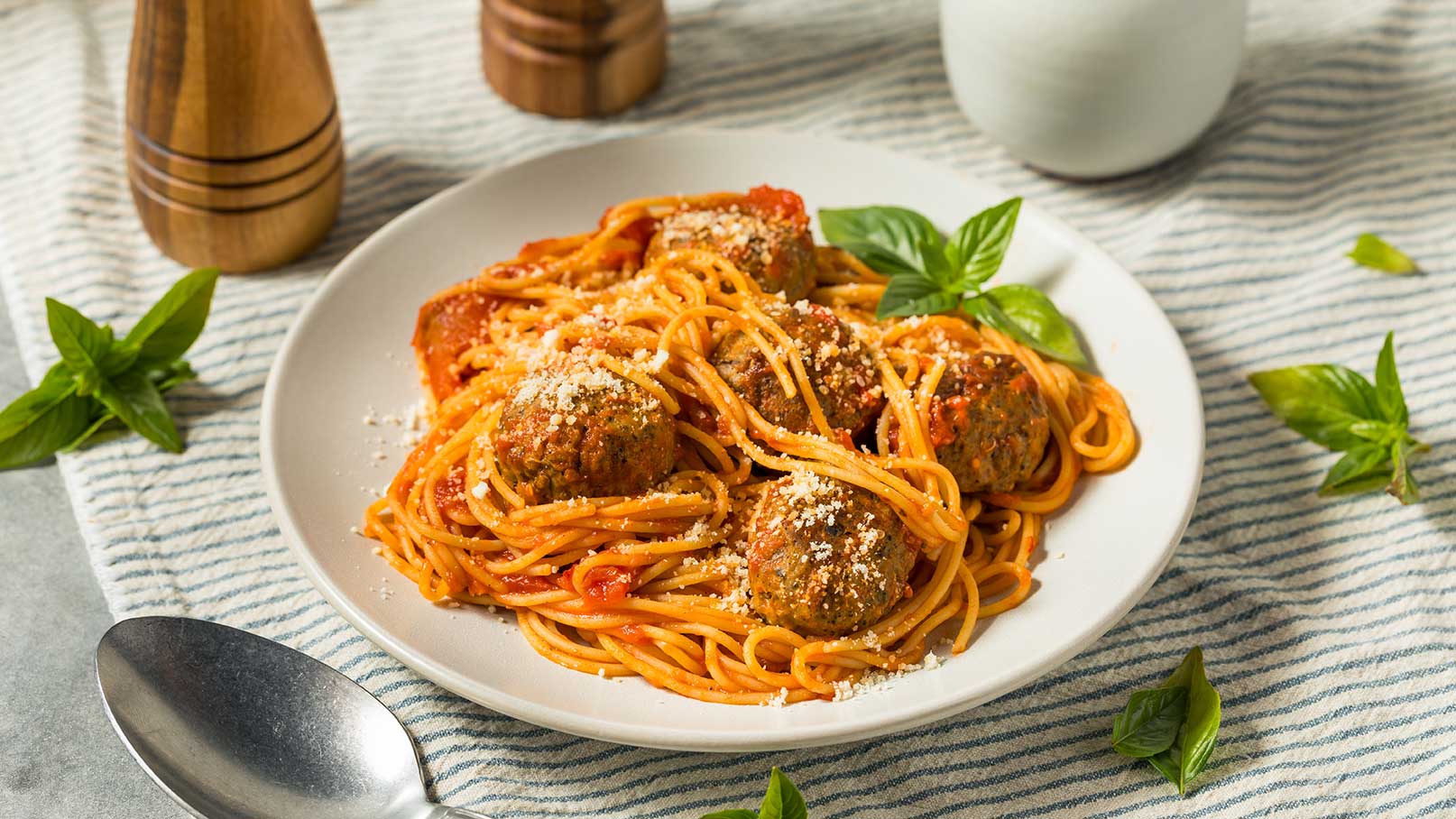 Ein Teller mit Spaghetti in Tomatensauce, garniert mit Käse und Basilikum. Die Spaghetti sind mit Fleischbällchen serviert und auf einem gestreiften Tischläufer platziert. Ein Löffel liegt neben dem Teller.