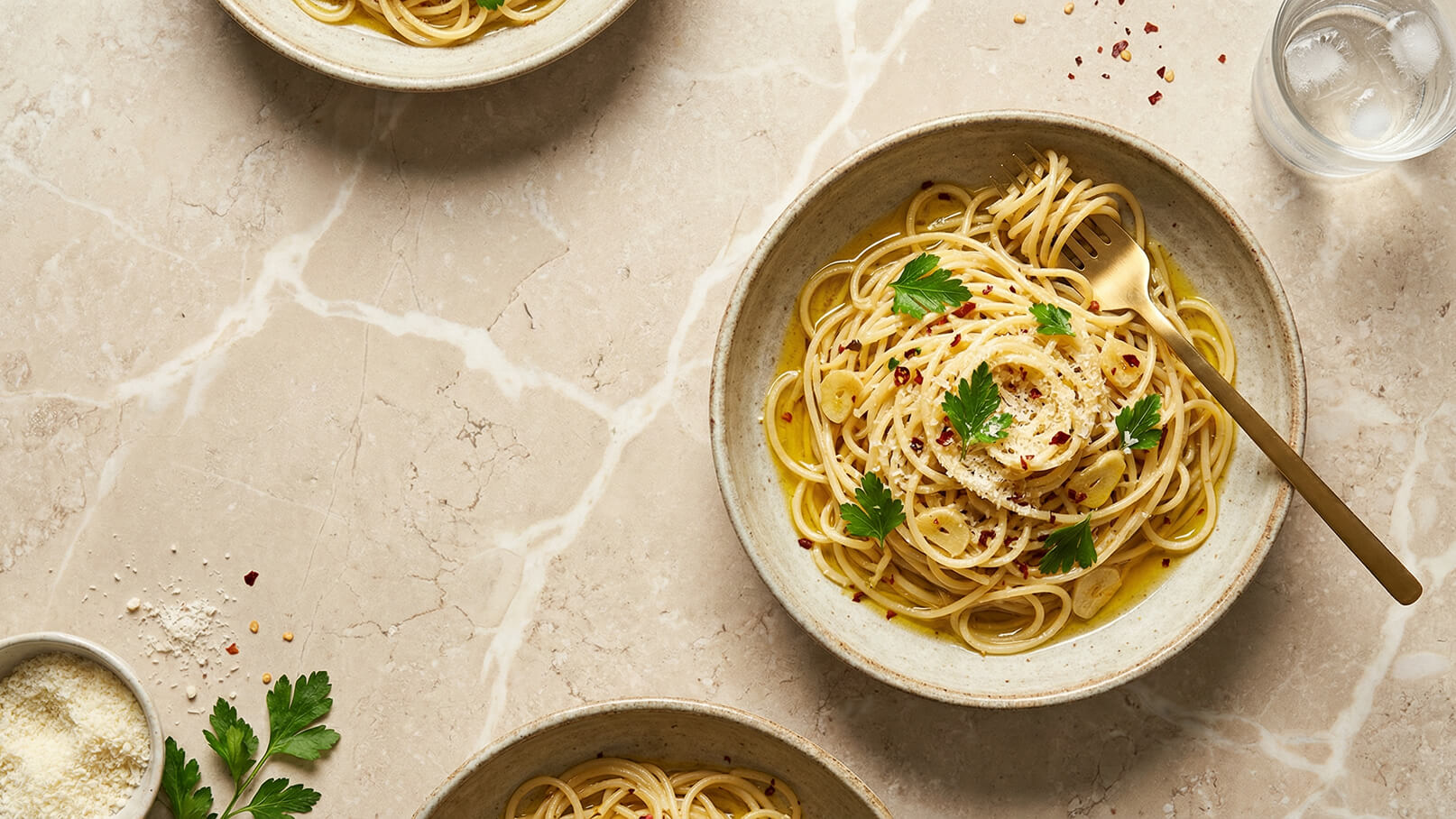 Ein Teller Spaghetti mit Olivenöl, Knoblauchscheiben und roten Pfefferflocken, garniert mit frischer Petersilie. Der Teller ist auf einer marmorierten Oberfläche platziert, daneben befindet sich ein weiteres kleines Gericht und ein Glas Wasser.