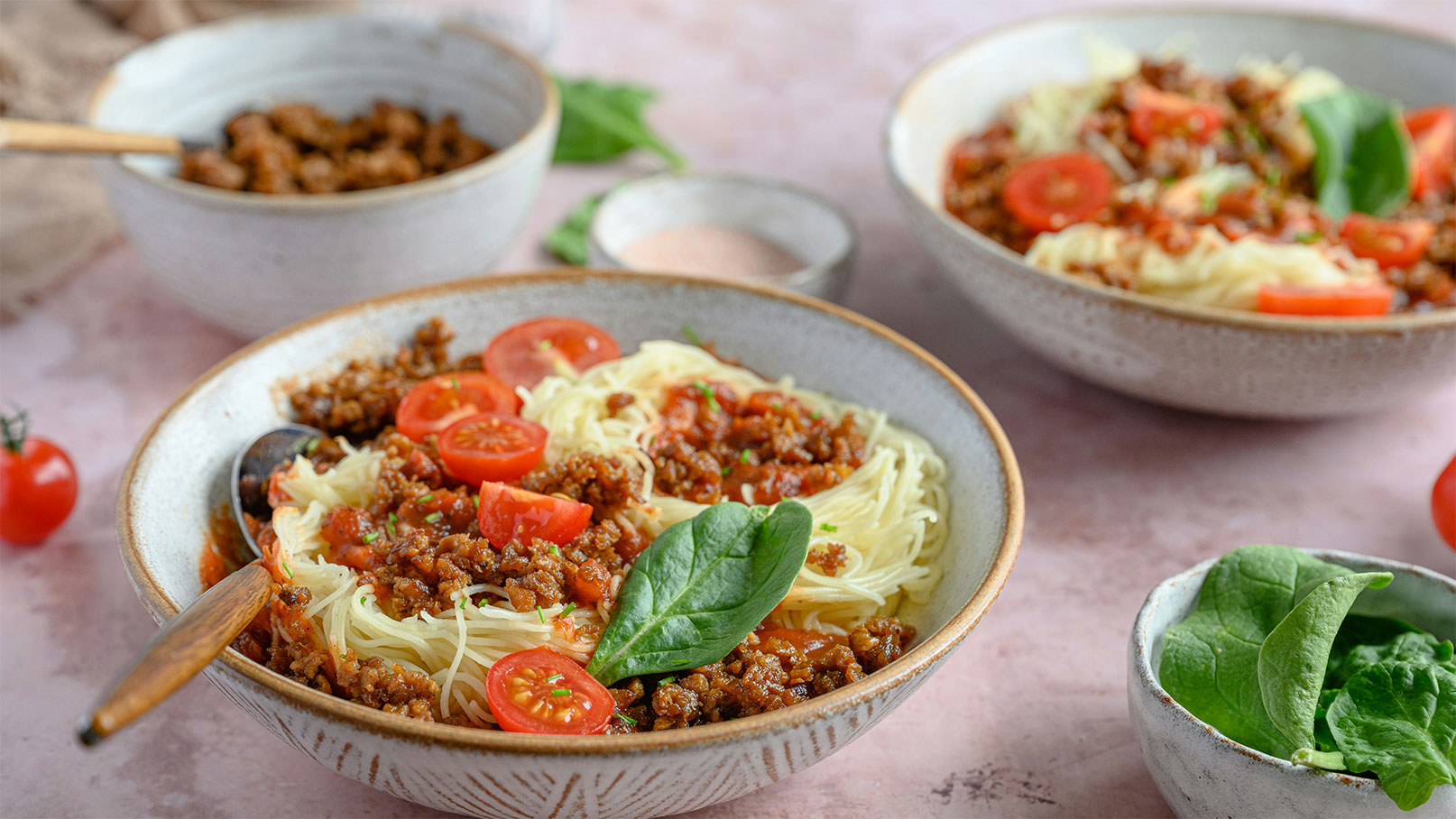 Zwei Schalen mit Spaghetti, überzogen mit einer würzigen Hackfleischsauce, sind im Vordergrund zu sehen. Frische Kirschtomaten und ein Blatt Spinat dekorieren die Pasta. Im Hintergrund gibt es eine zusätzliche Schale mit Hackfleisch und eine kleine Schale mit Salz.