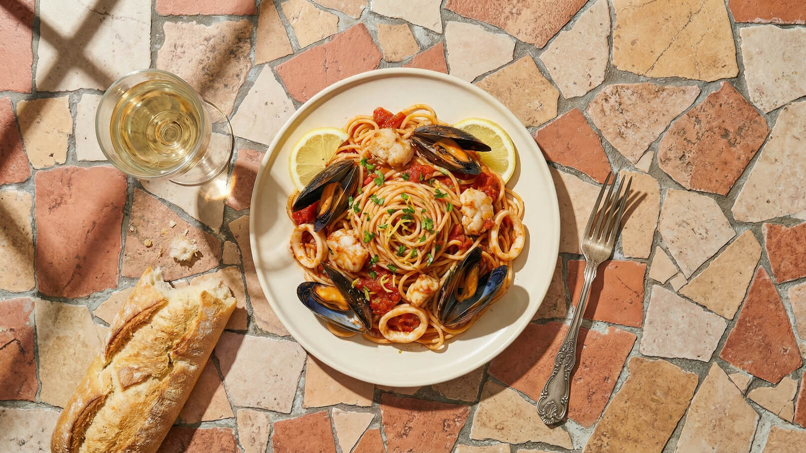 Ein Teller Pasta mit Meeresfrüchten, serviert in einer Tomatensauce. Er ist garniert mit frischen Zitronenscheiben und gehacktem Grün. Daneben steht ein Glas Weißwein und ein Stück Baguette liegt auf dem Tisch aus bunten Steinfliesen.