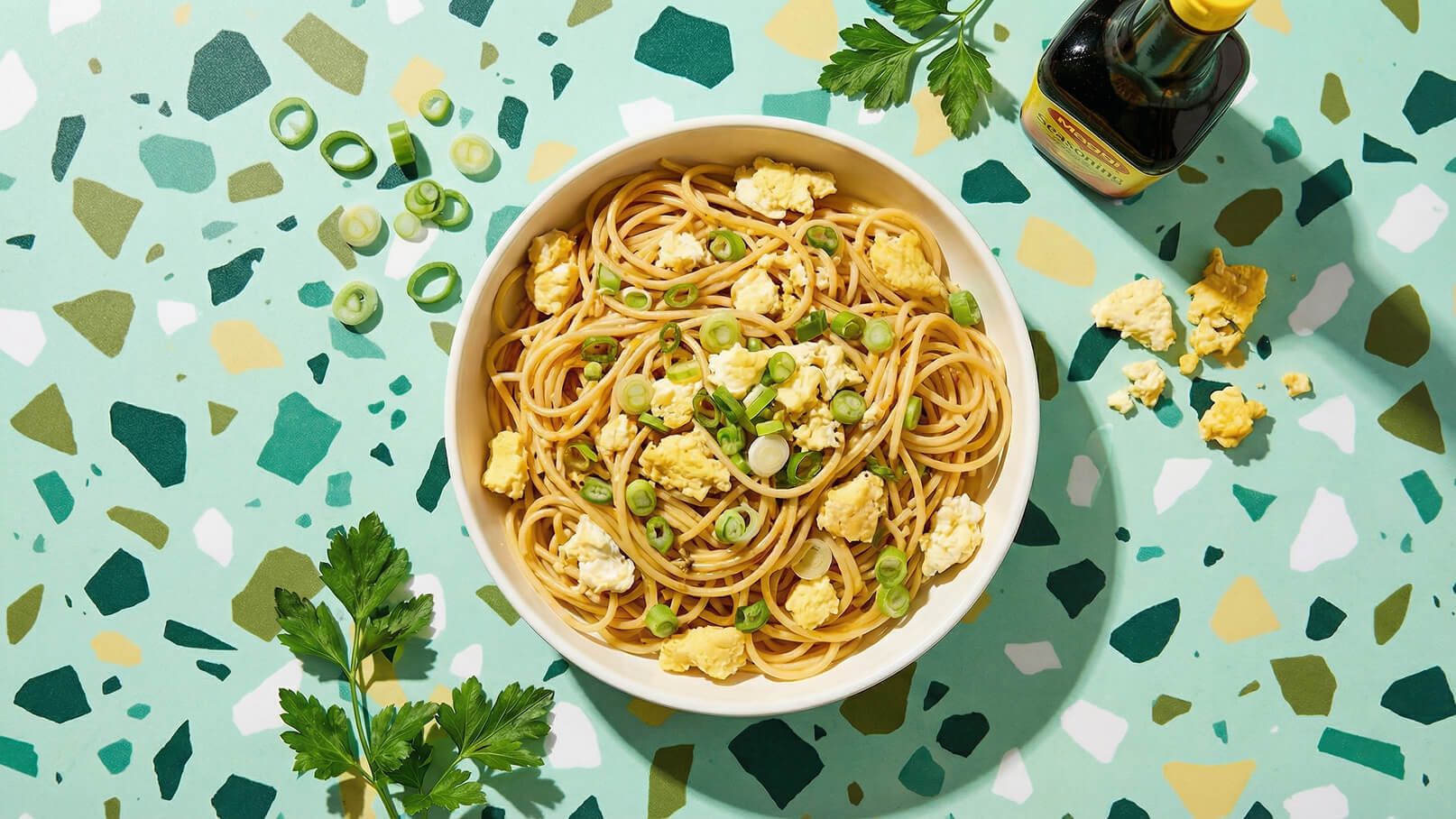 Eine Schüssel mit Spaghetti, garniert mit Rührei und Frühlingszwiebeln, steht auf einem bunten, strukturierten Untergrund. Neben der Schüssel liegt ein Bund frischer Petersilie und eine Flasche Sojasauce.