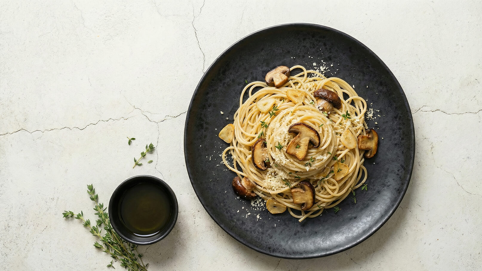Ein schwarzer Teller mit Spaghetti, garniert mit gebratenen Pilzen, gehacktem Knoblauch und frisch geriebenem Käse. Daneben steht eine kleine Schüssel mit Olivenöl und einige Kräuter liegen auf der Oberfläche.