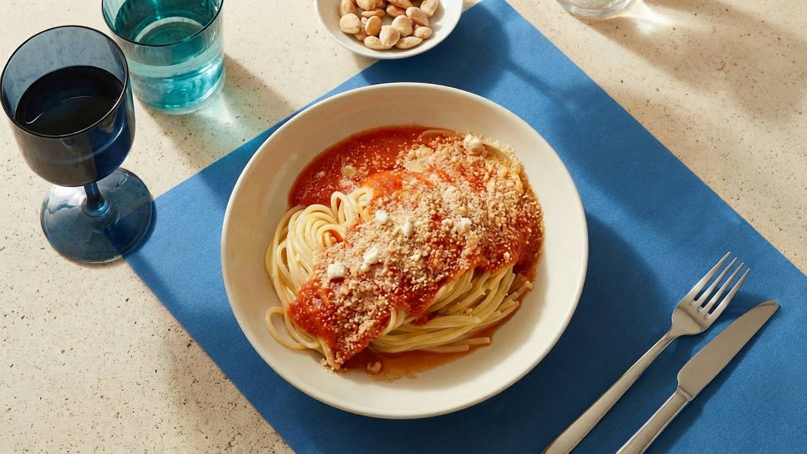 Ein Teller mit Spaghetti, bedeckt mit Tomatensoße und Käse, steht auf einem blauen Tischset. Daneben befinden sich ein glas Wasser und ein Glas Wein, sowie eine Schale mit Mandeln. Besteck ist ordentlich angelegt.