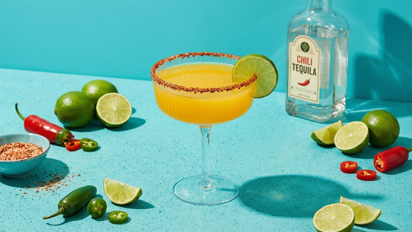 Ein Cocktailglas mit einer orangefarbenen Flüssigkeit, garniert mit einem Limettenschnitz und einem Zuckerrand, steht auf einem türkisfarbenen Tisch. Um das Glas herum liegen verschiedene Limetten und Chillischoten. Eine Flasche mit der Aufschrift „Chili Tequila“ ist im Hintergrund sichtbar.