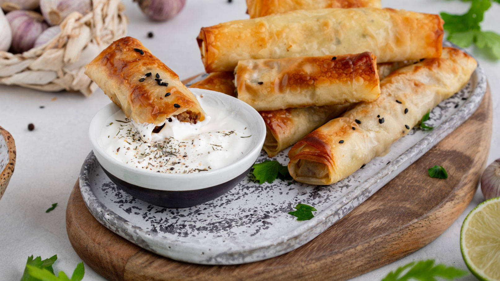 Ein Teller mit knusprigen Röllchen aus Filoteig, gefüllt mit Fleisch, liegt auf einer rustikalen Holzplatte. Daneben befindet sich eine kleine Schüssel mit Joghurt-Dip, garniert mit Gewürzen. Im Hintergrund sind einige Küchenelemente, wie Knoblauch und frische Kräuter, sichtbar.
