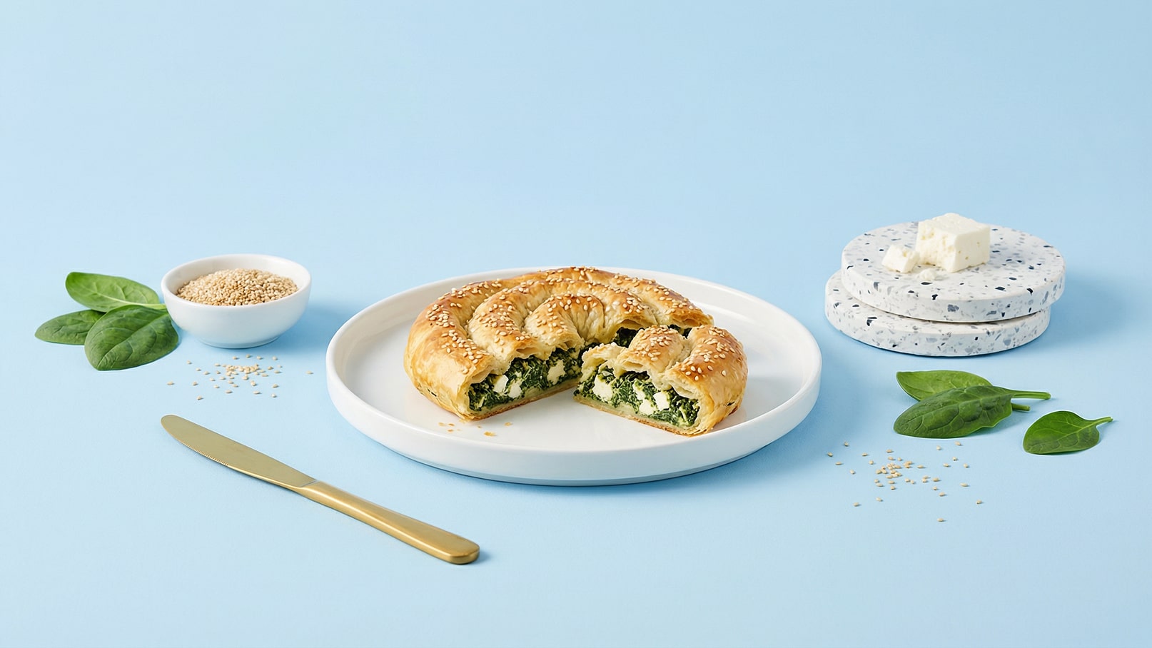 Ein Teller mit einem halbierten gefüllten Blätterteiggebäck, das mit Sesam bestreut ist. Die Füllung besteht aus Spinat und Käse. Neben dem Teller liegen einige frische Spinatblätter, Sesam und ein goldener Messer. Im Hintergrund sind zwei Untersetzer mit Käse.