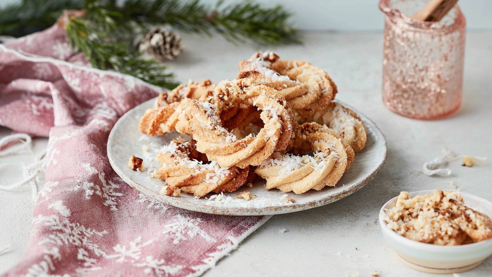 Auf einem dunklen Hintergrund liegen verschiedene Weihnachtsplätzchen, darunter geformte Kekse und einige rechteckige. Getrocknete Orangenscheiben und Tannenzweige sind ebenfalls sichtbar, während Puderzucker verstreut ist und eine Sterneform hervorgehoben wird.