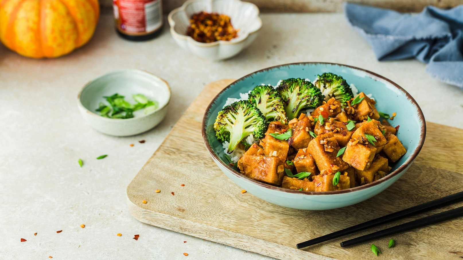 Eine Schüssel mit gebratenem Tofu auf Reis, garniert mit Brokkoli und grünen Zwiebeln. Der Tofu ist in einer würzigen Sauce mariniert. Im Hintergrund befinden sich kleine Schalen mit Frischhalte- und Chilipulver. Holzbrett und Essstäbchen sind sichtbar.