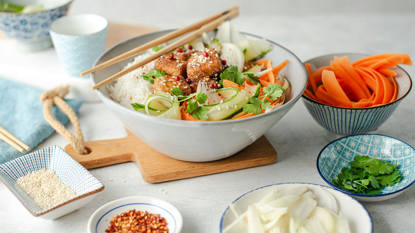 Eine Schüssel mit Reis, garniert mit gebratenem Tofu, frischem Gemüse, wie Karotten und Gurken, sowie Koriander. Die Schüssel ist auf einem Holzbrett platziert und umgeben von kleinen Schalen mit Sesam und roten Pfefferflocken. Essstäbchen liegen bereit zum Essen.