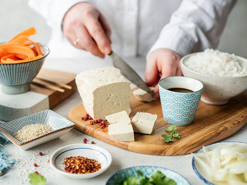 Eine Person schneidet Tofu auf einem Holzbrett. Neben dem Tofu befinden sich Schalen mit Gewürzen und eine Tasse. Im Hintergrund ist eine Schüssel Reis sichtbar. Die Szene vermittelt eine Vorbereitungsphase zum Kochen.