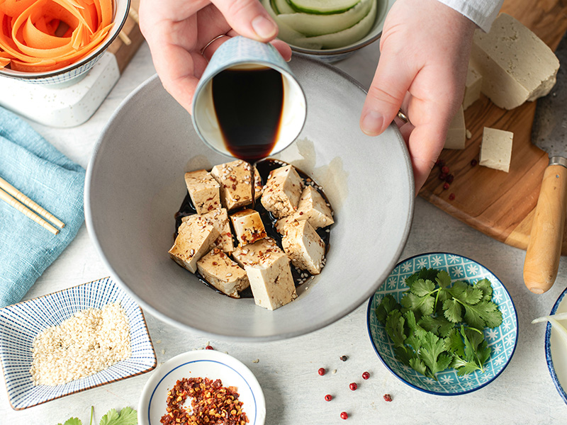 Eine Hand gießt Sojasauce über würfelförmigen Tofu in einer großen Schüssel. Der Tofu ist mit Sesam und Chiliflocken bestreut. Im Hintergrund sind verschiedene Zutaten wie frischer Koriander und andere Gewürze sichtbar.