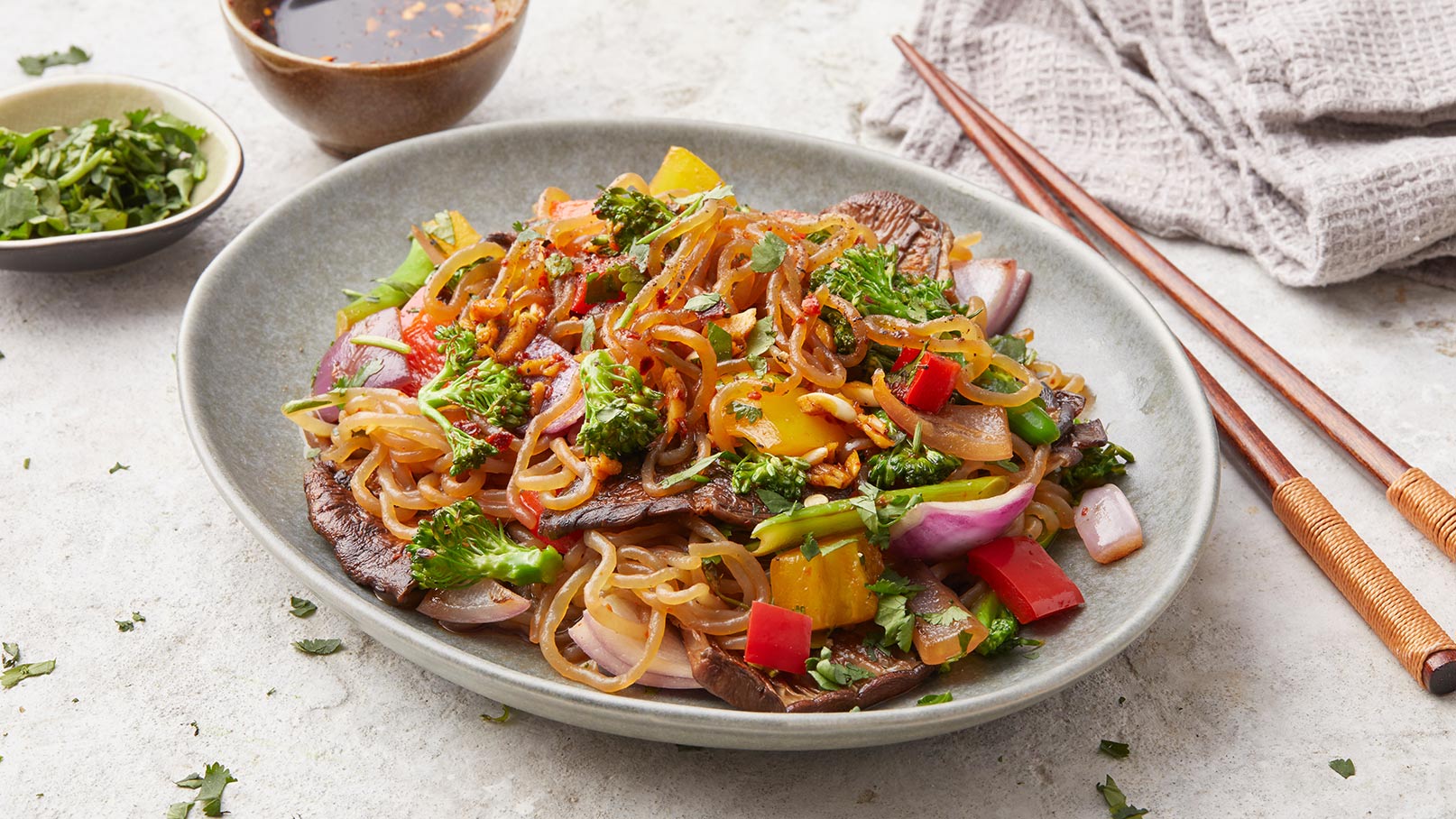 Ein Teller mit buntem gebratenem Nudelgericht, das Gemüse wie Paprika, Brokkoli und Zwiebeln enthält. Das Gericht ist mit frischen Kräutern garniert. Im Hintergrund sind Essstäbchen und eine Schale mit Sauce zu sehen.