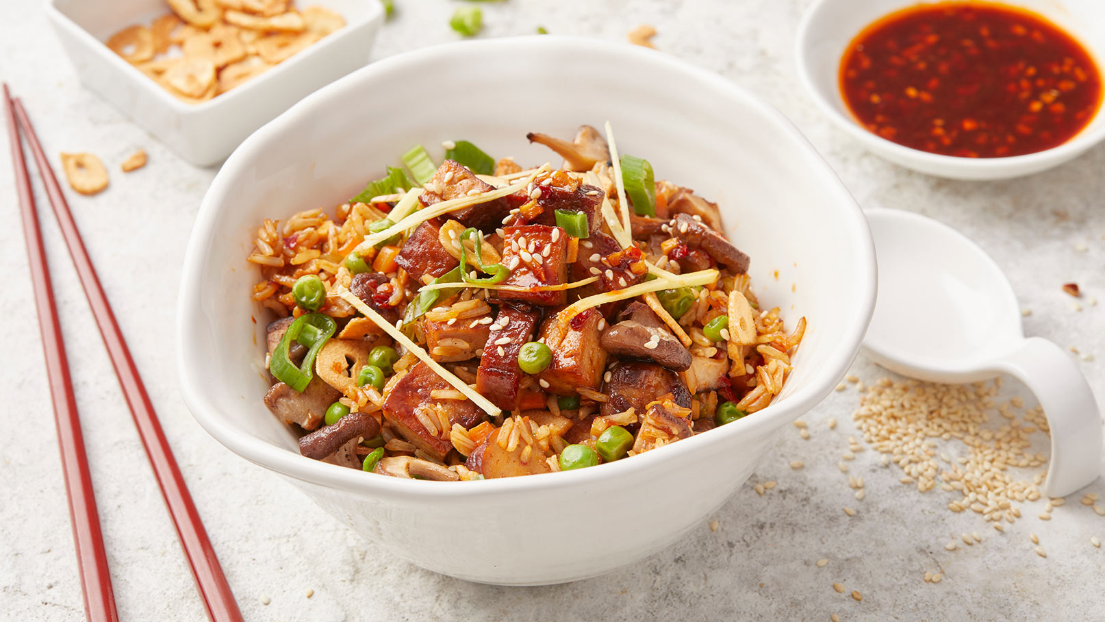 Eine weiße Schüssel mit gebratenem Reis, Stückchen von Gemüse und Fleisch, garniert mit Frühlingszwiebeln und Ingwerstreifen. Neben der Schüssel liegen Essstäbchen und es gibt eine kleine Schale mit roter Sauce. Der Hintergrund ist hell und schlicht.